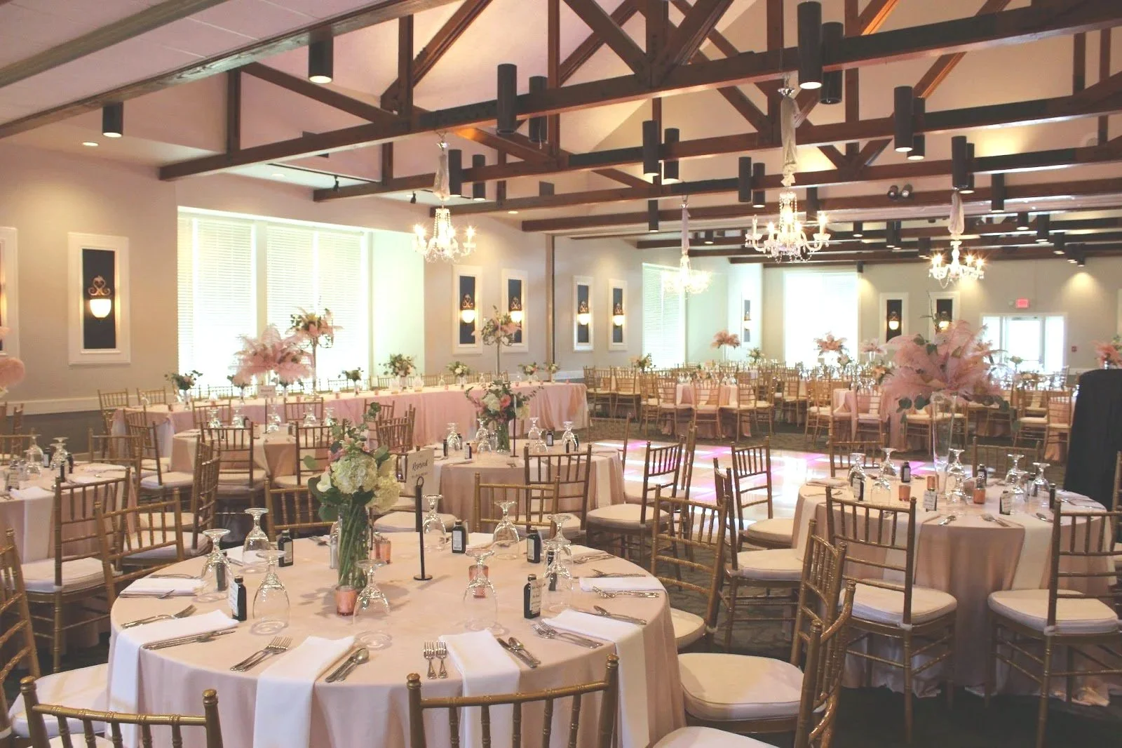 Wildwood CC round tables, cream tablecloths, and gold Chiavari chairs. Tall floral centerpieces Chandeliers hang from a wooden-beam ceiling, and wall sconces provide soft lighting.