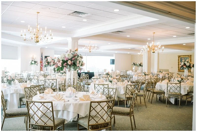 Elegant banquet hall set up for a wedding reception with round tables, floral centerpieces, gold chairs, chandeliers, and soft lighting.