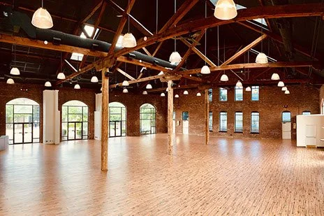 Spacious empty brick-walled room with large windows, wooden beams, and hanging lights.