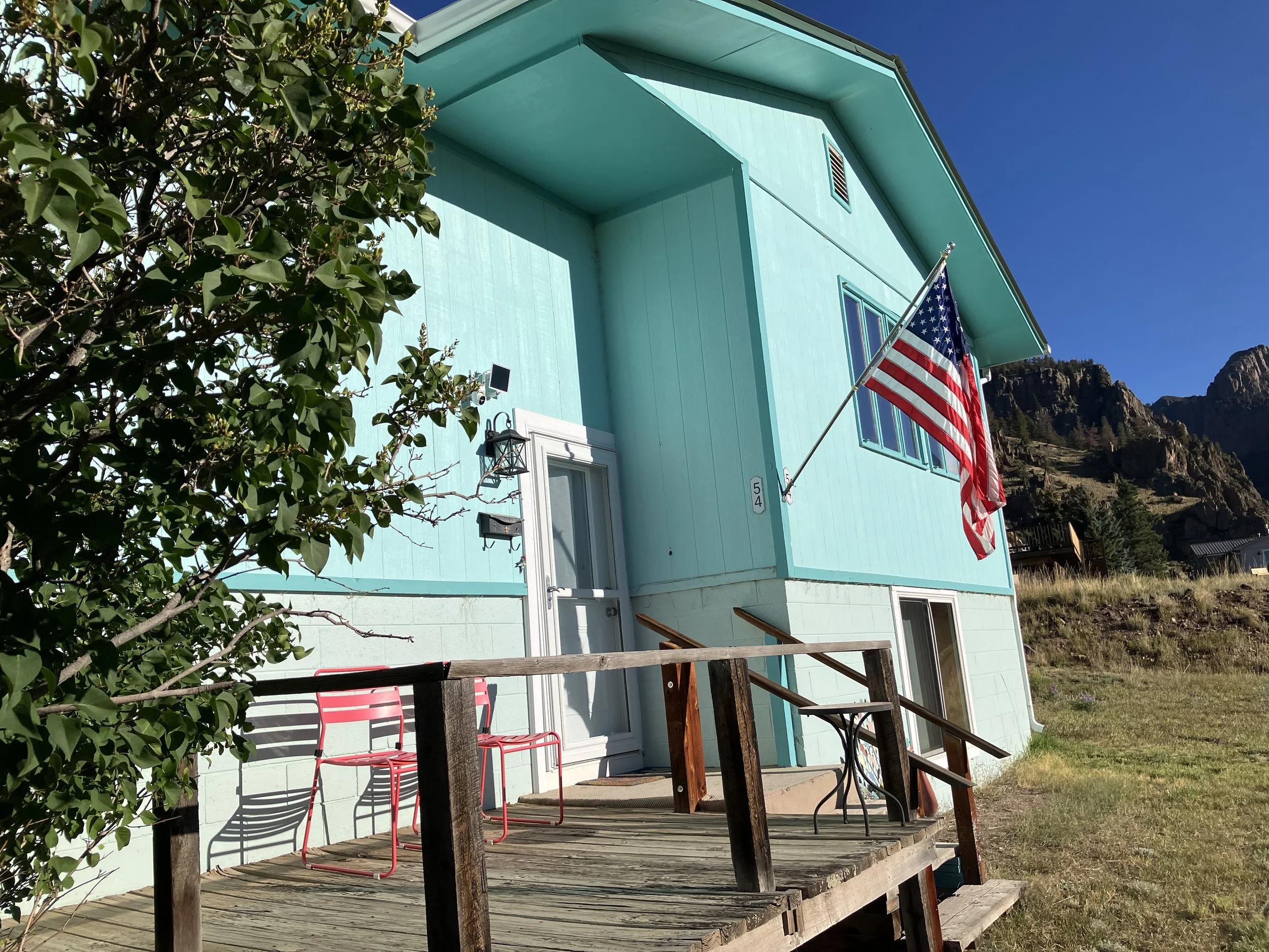 A light blue house with a wooden deck, two pink chairs, and an American flag hanging outside. There are mountains in the background under a clear blue sky.
