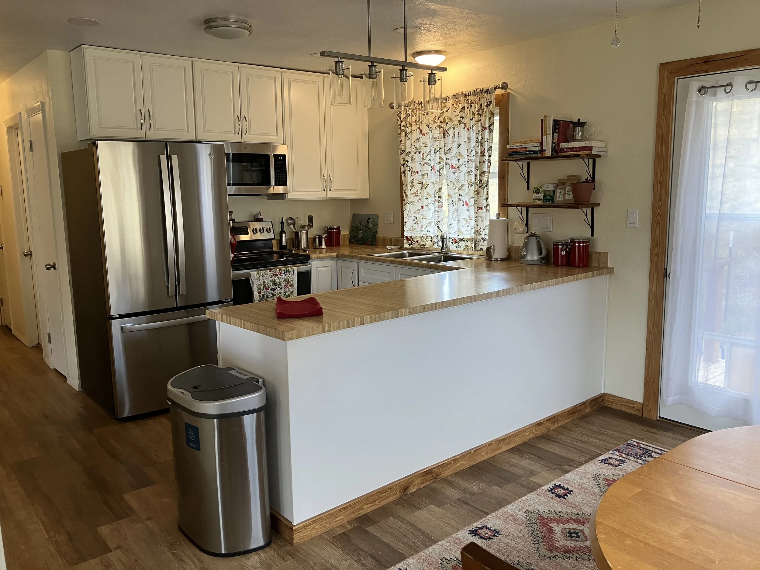 Kitchen with white cabinets, stainless steel refrigerator, microwave, and oven, wooden countertops, a window with floral curtain, shelves with books and kitchen items, a paper towel holder, and a sliding door with sheer curtain.