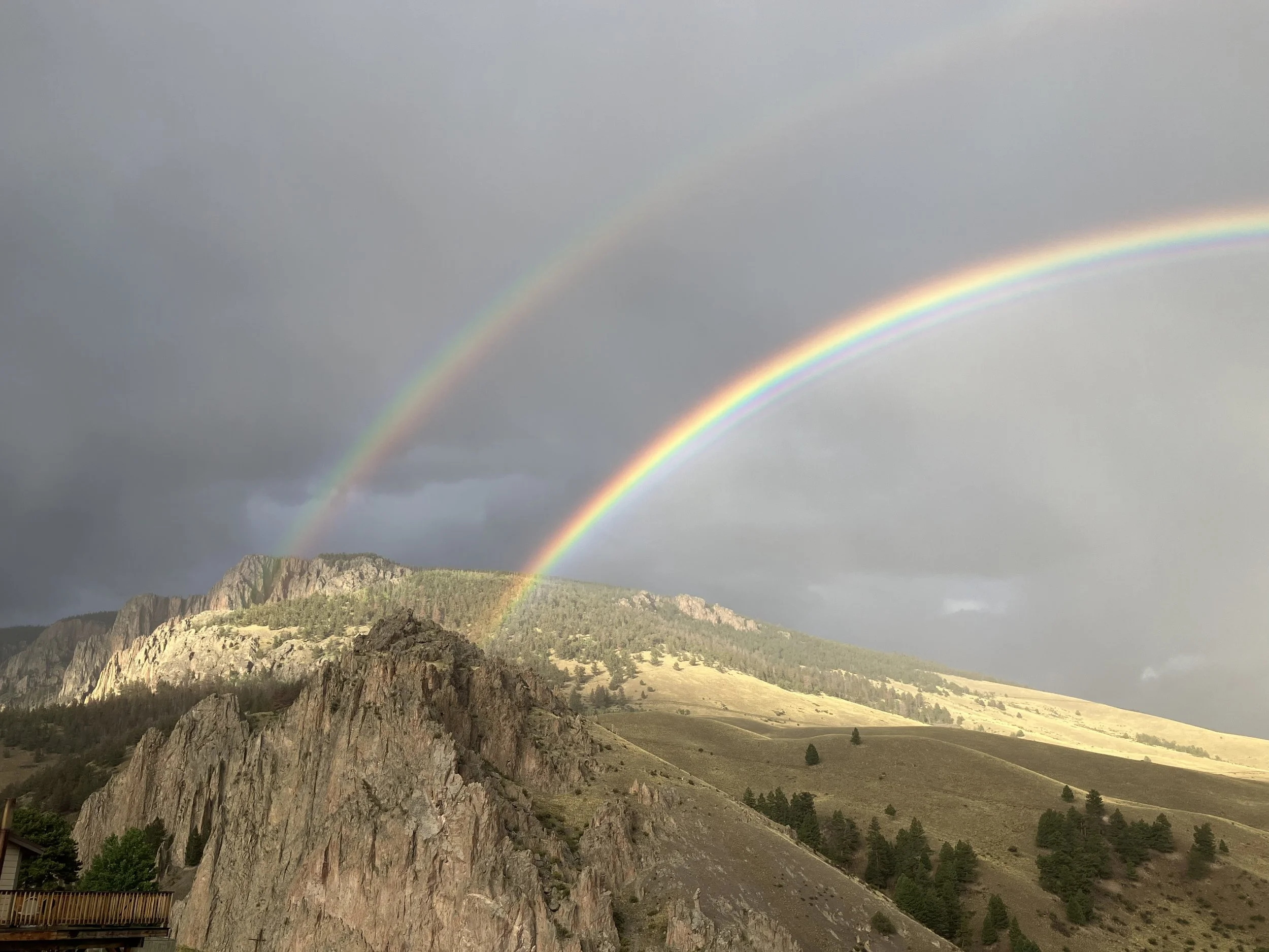Two rainbows arching over a mountainous landscape with rocky cliffs, grassy slopes, and scattered trees under a cloudy sky.
