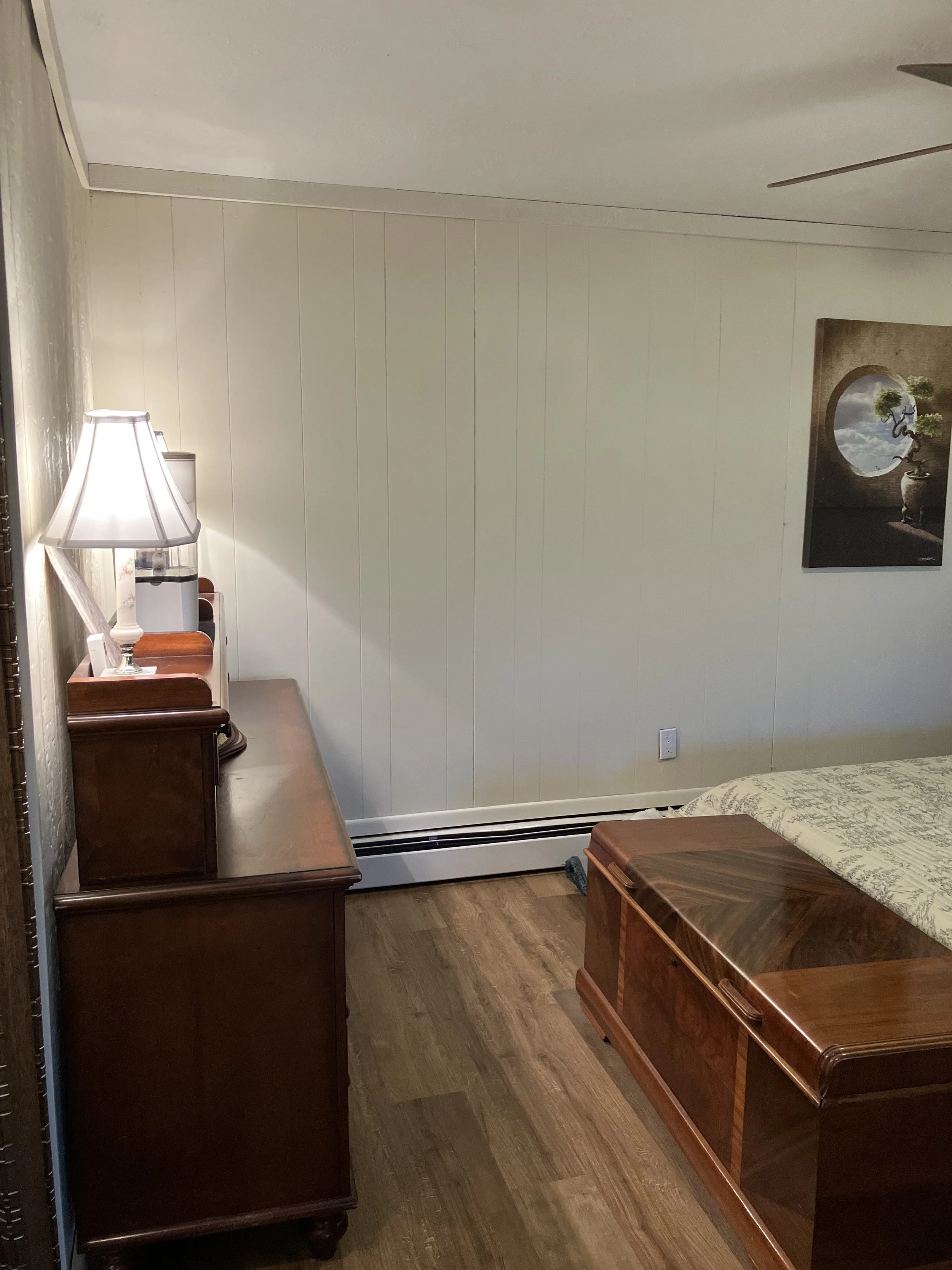 A bedroom corner with a wooden dresser with a lamp and a white water dispenser on top, a bed with a wooden headboard, a partially visible painting of a moonlit landscape, and a wall-mounted electrical outlet.