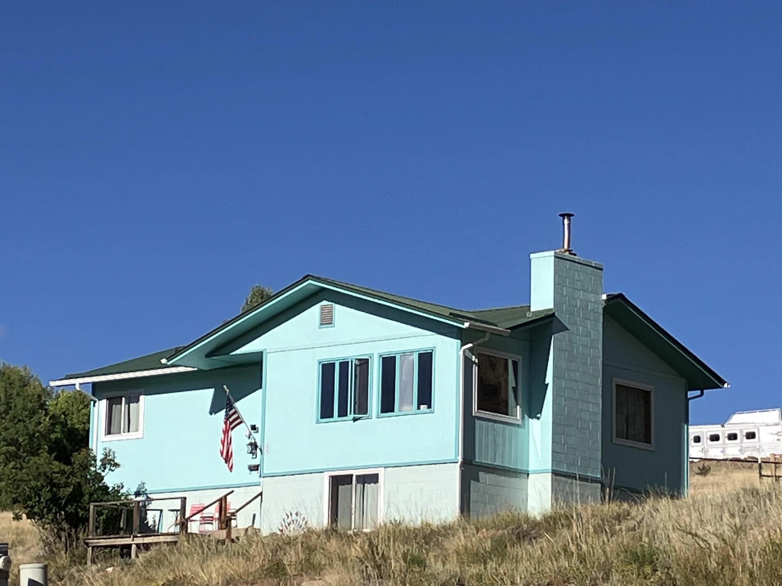 A turquoise house on a grassy hill under a clear blue sky, with an American flag hanging from a pole on the front porch.