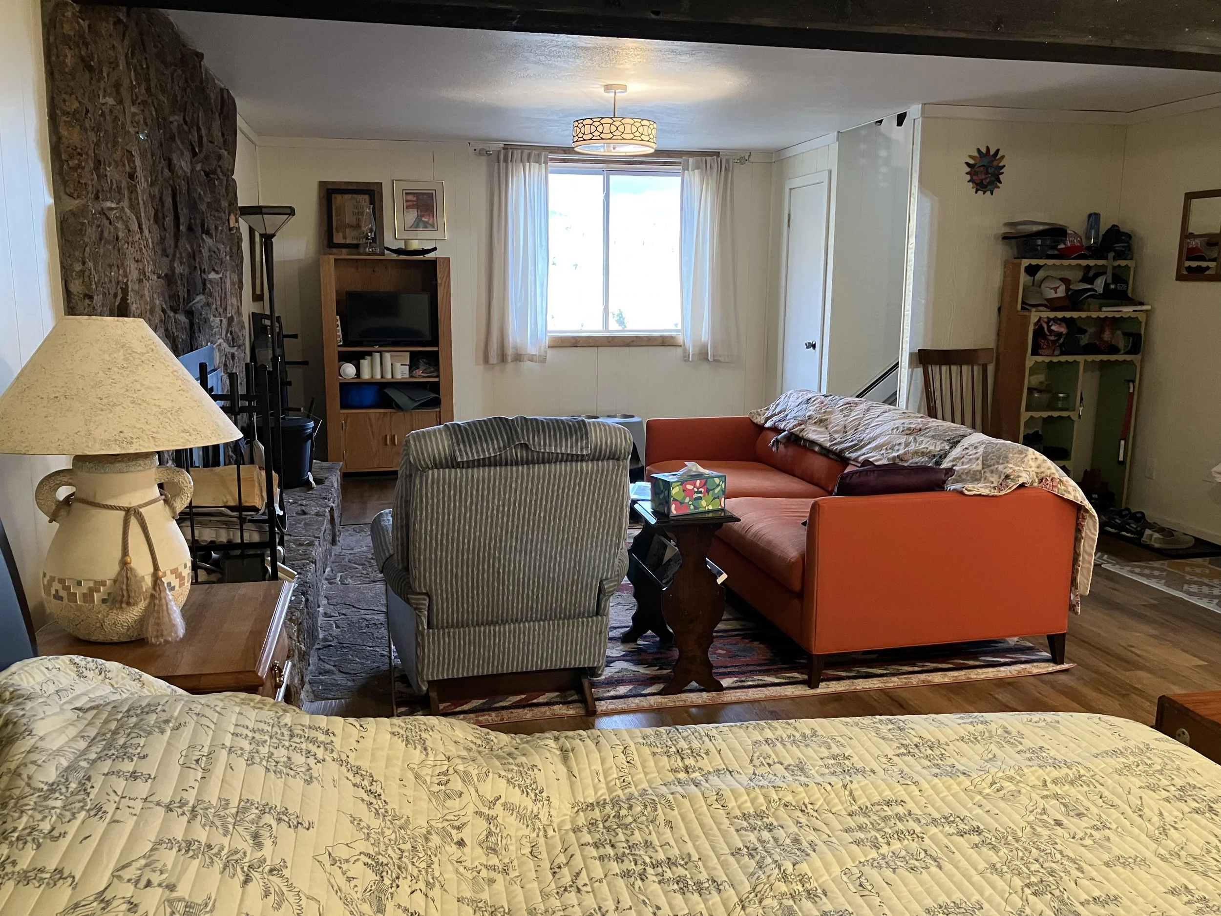 Living room with a bed in the foreground, a stone fireplace on the left, a beige armchair, a bright orange couch with a blanket, a window with curtains, a TV on a wooden stand, and various decorative items and furniture.