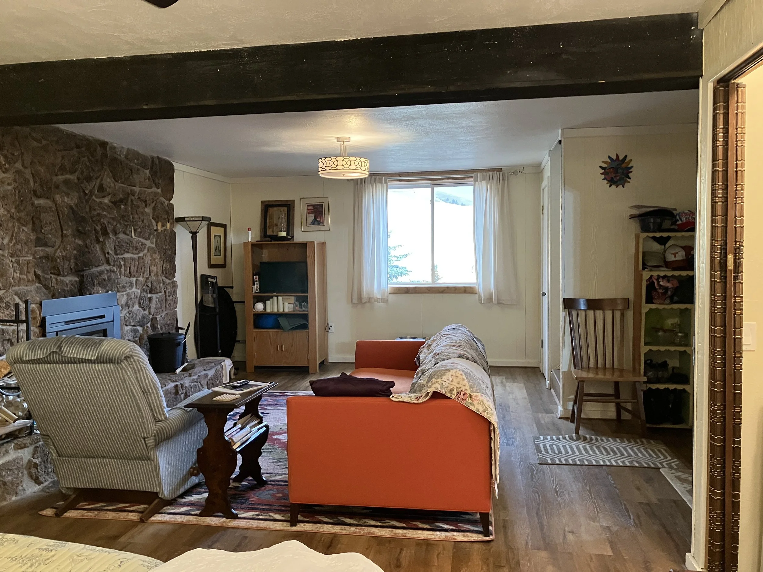 Living room with a stone fireplace, orange couch, armchair, wooden side table, and a window with white curtains, decorated with family photos and various items on shelves.