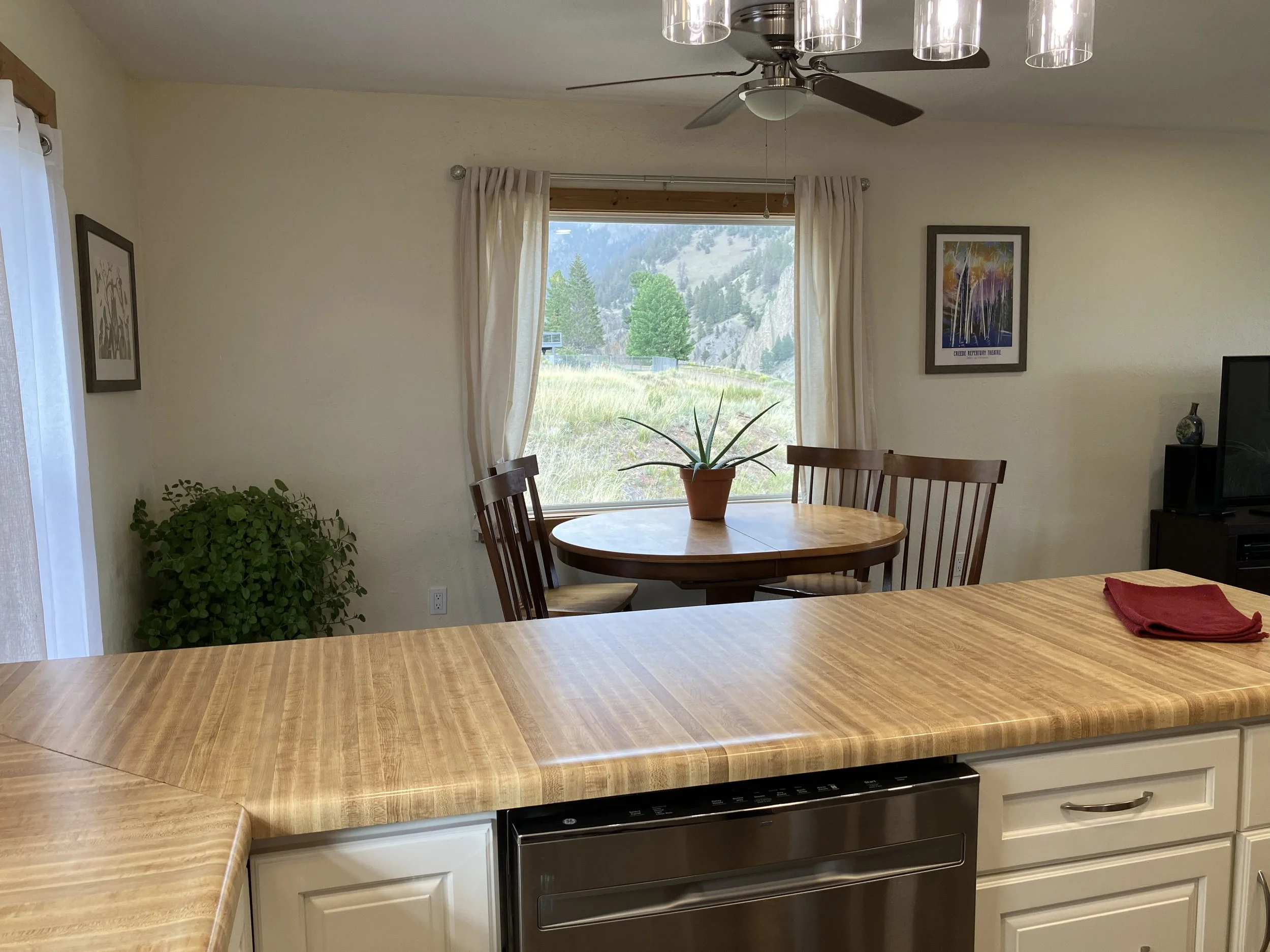 View of a kitchen with a wooden countertop, a dining area with a round table, three wooden chairs, a window with curtains showing a scenic outdoor landscape, a potted plant on the table, and framed artwork on the walls.