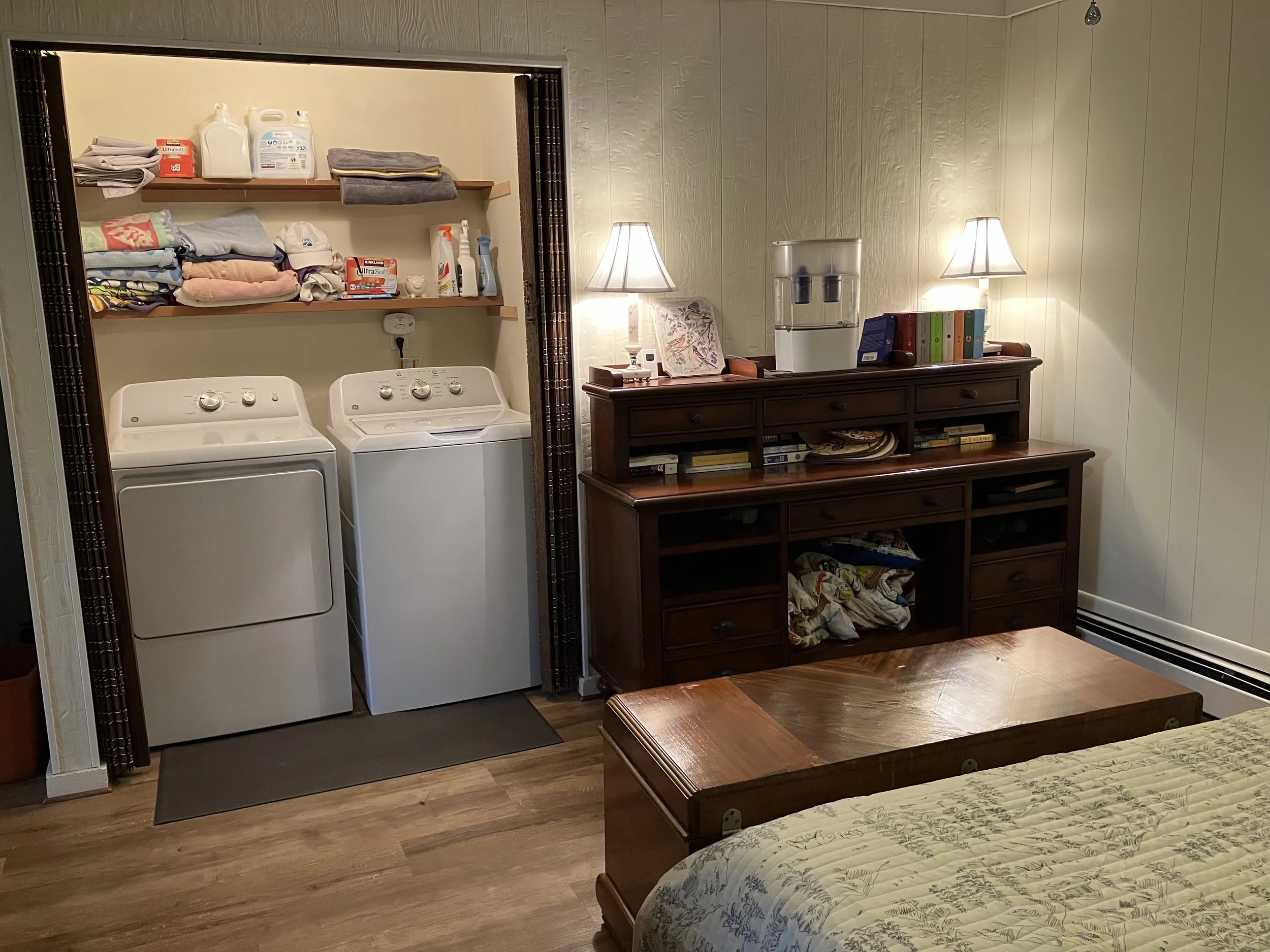 Laundry area with a top-loading washer and dryer, shelves with laundry supplies and towels, next to a wooden dresser with books, a water dispenser, and lamps in a cozy bedroom.