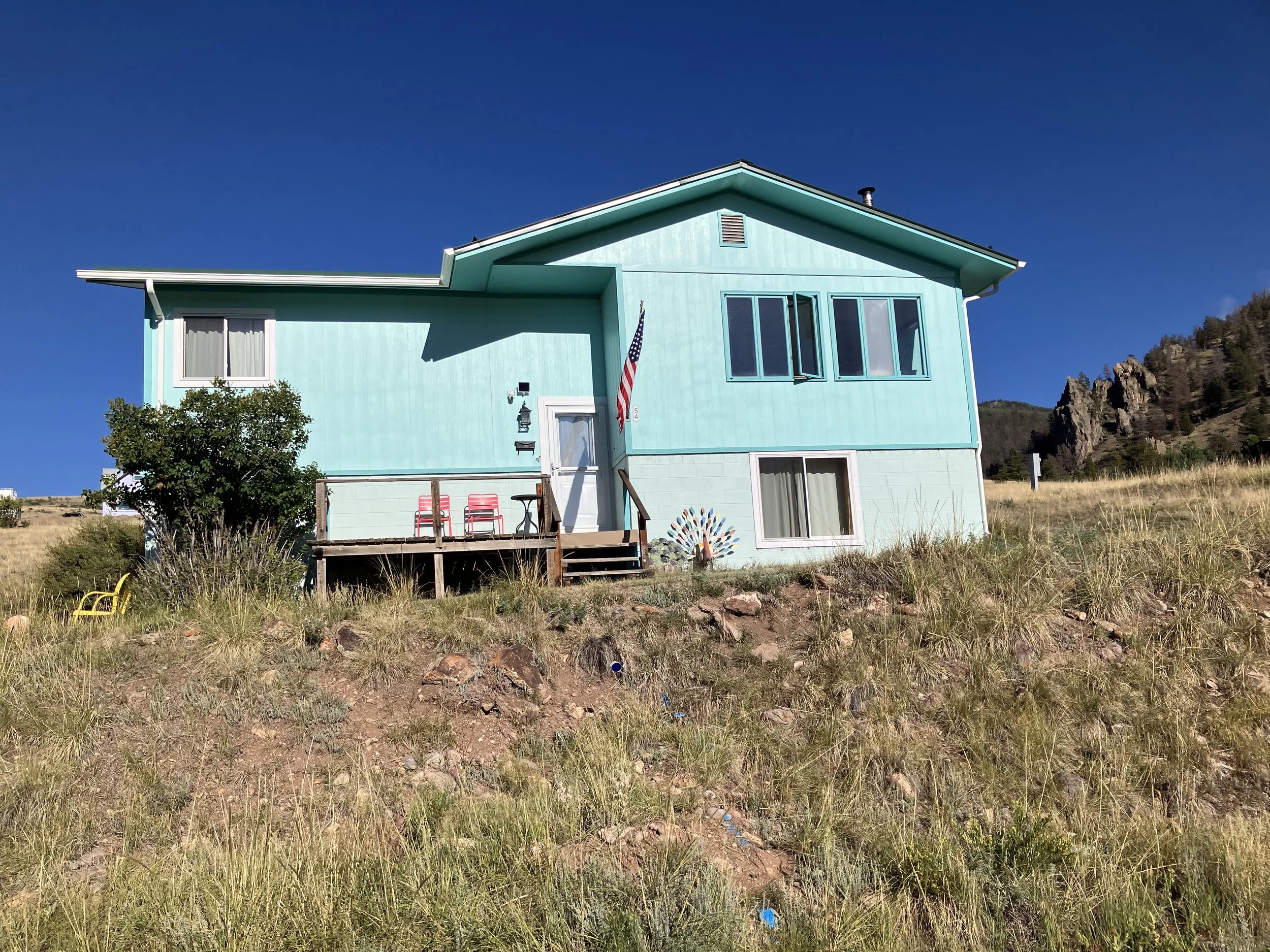 A two-story turquoise house with a small front porch, set on a grassy hillside with rocky terrain, against a clear blue sky. The house has large windows and an American flag hanging near the door.