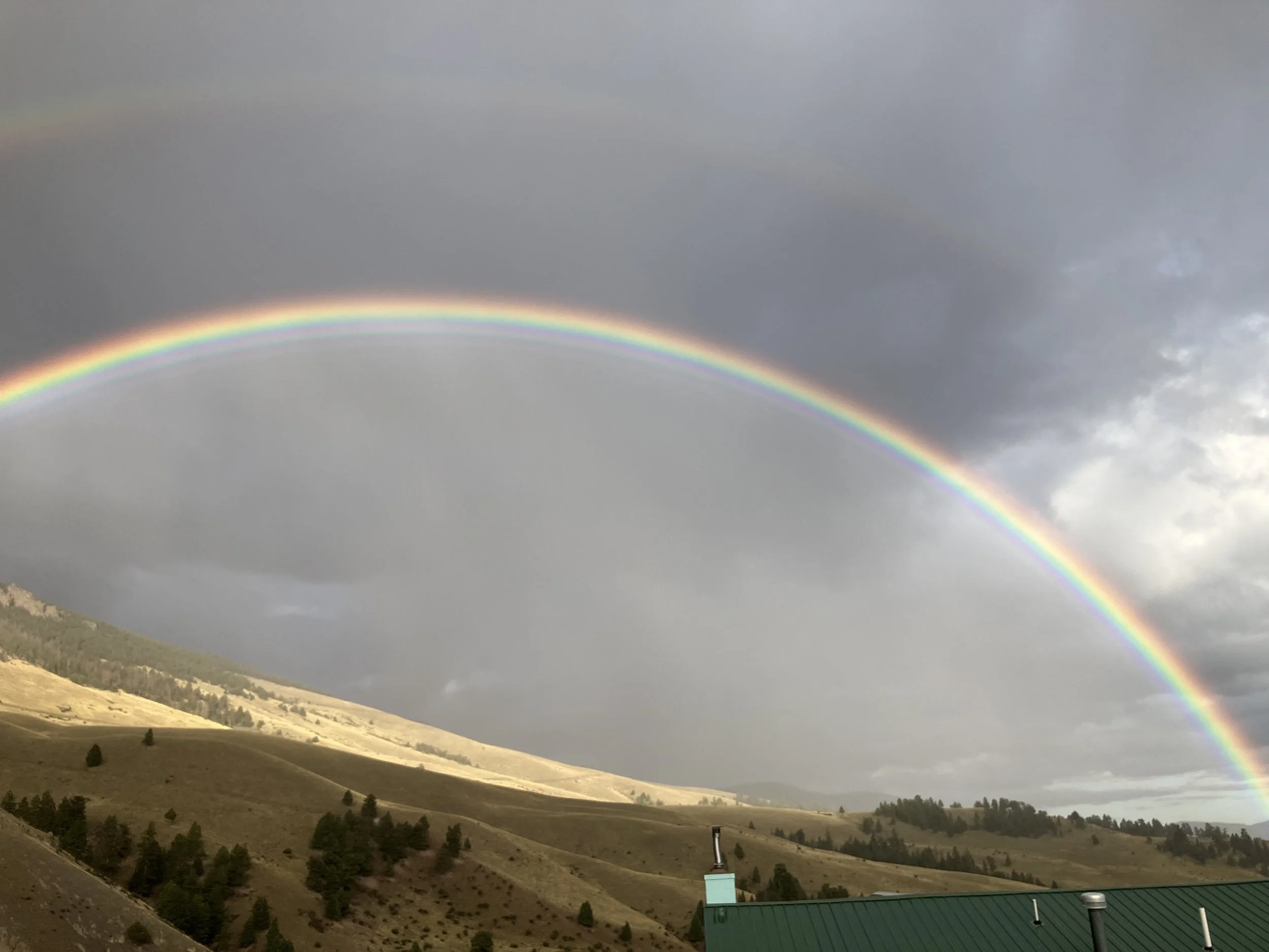A double rainbow arching over a hilly landscape with scattered trees and a building with a green roof.
