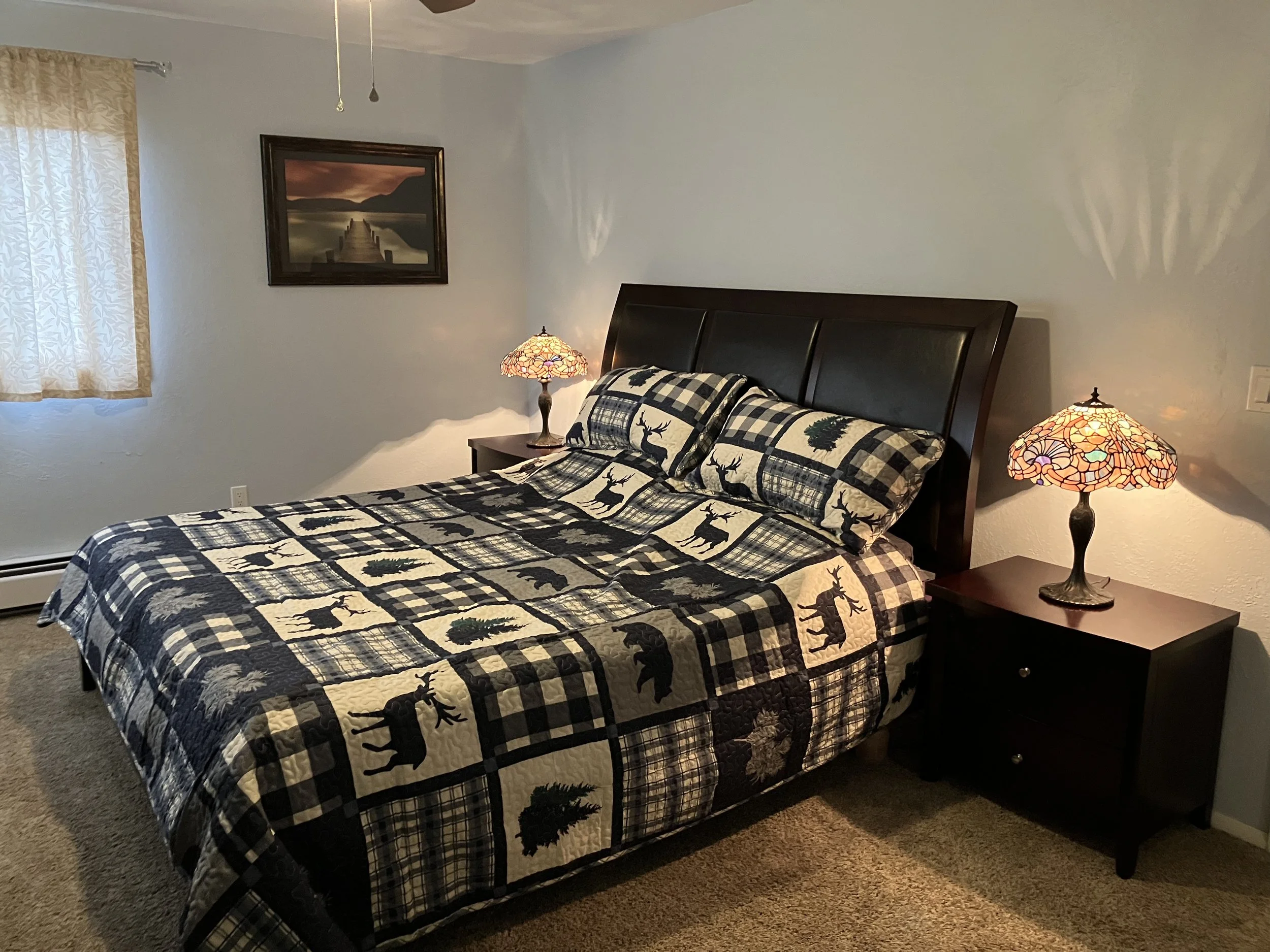 A bedroom with a bed covered in a holiday-themed quilt featuring deer and pine trees, and two matching pillows. There are two nightstands with stained glass lamps on each side of the bed. The room has a window with curtains and a framed landscape pic