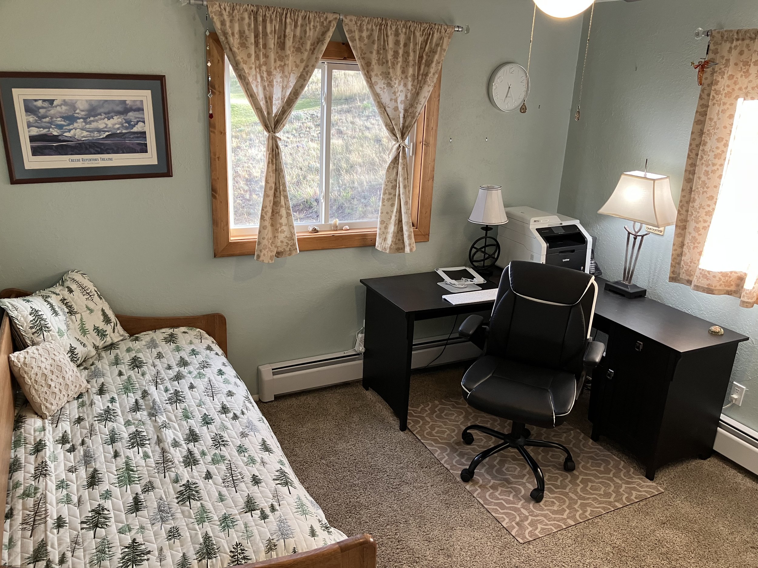 A bedroom with a bed covered in a pine tree patterned quilt on the left, a window with curtains in the center, and a black desk with a monitor, printer, desk lamp, and office chair on the right. There is a wall clock and framed picture above the bed,