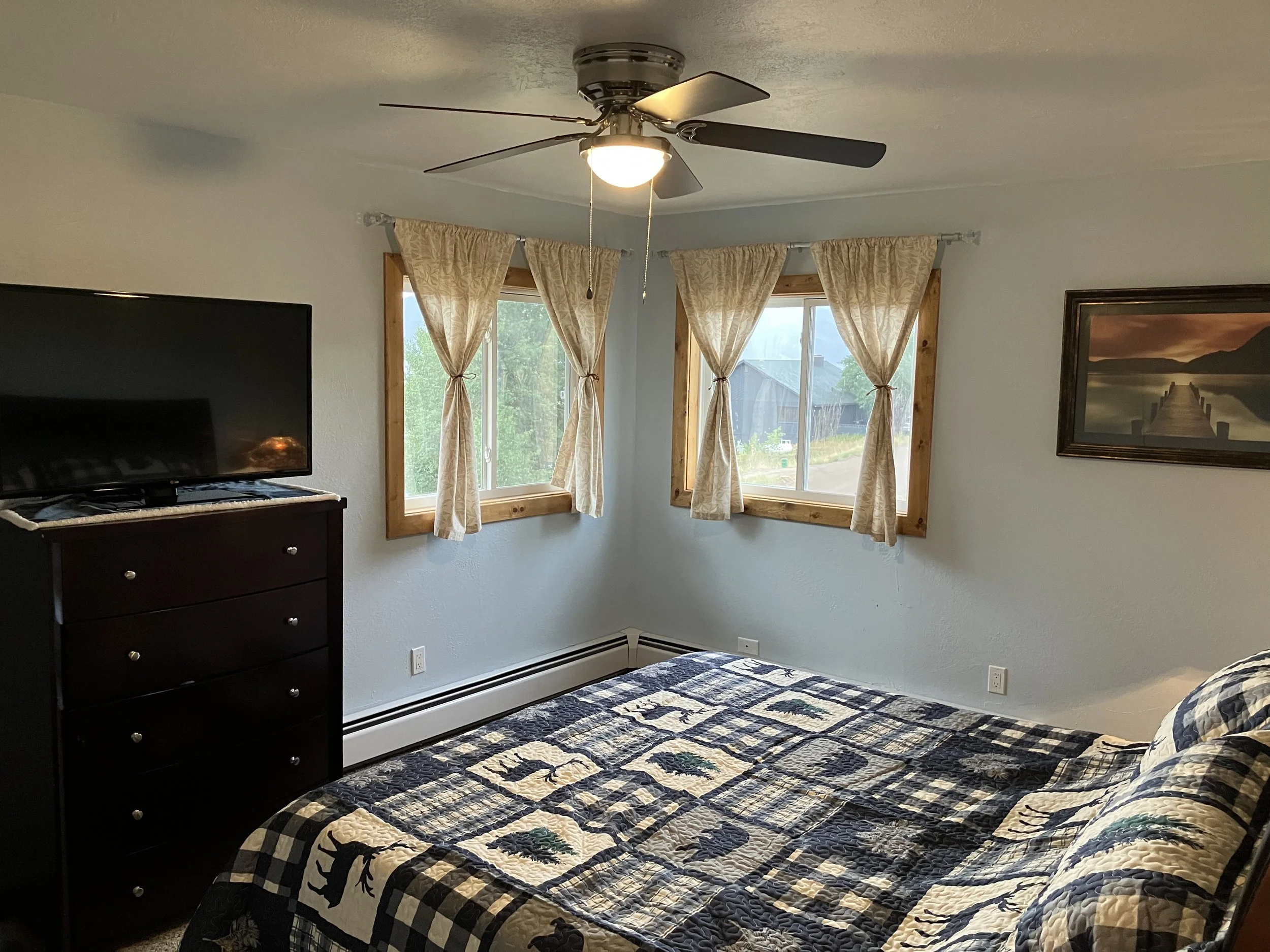 A bedroom with a ceiling fan, two windows with beige curtains, a dark wooden dresser with a TV on top, a framed landscape picture on the wall, and a bed with a quilted blanket featuring a rustic, nature-themed design.