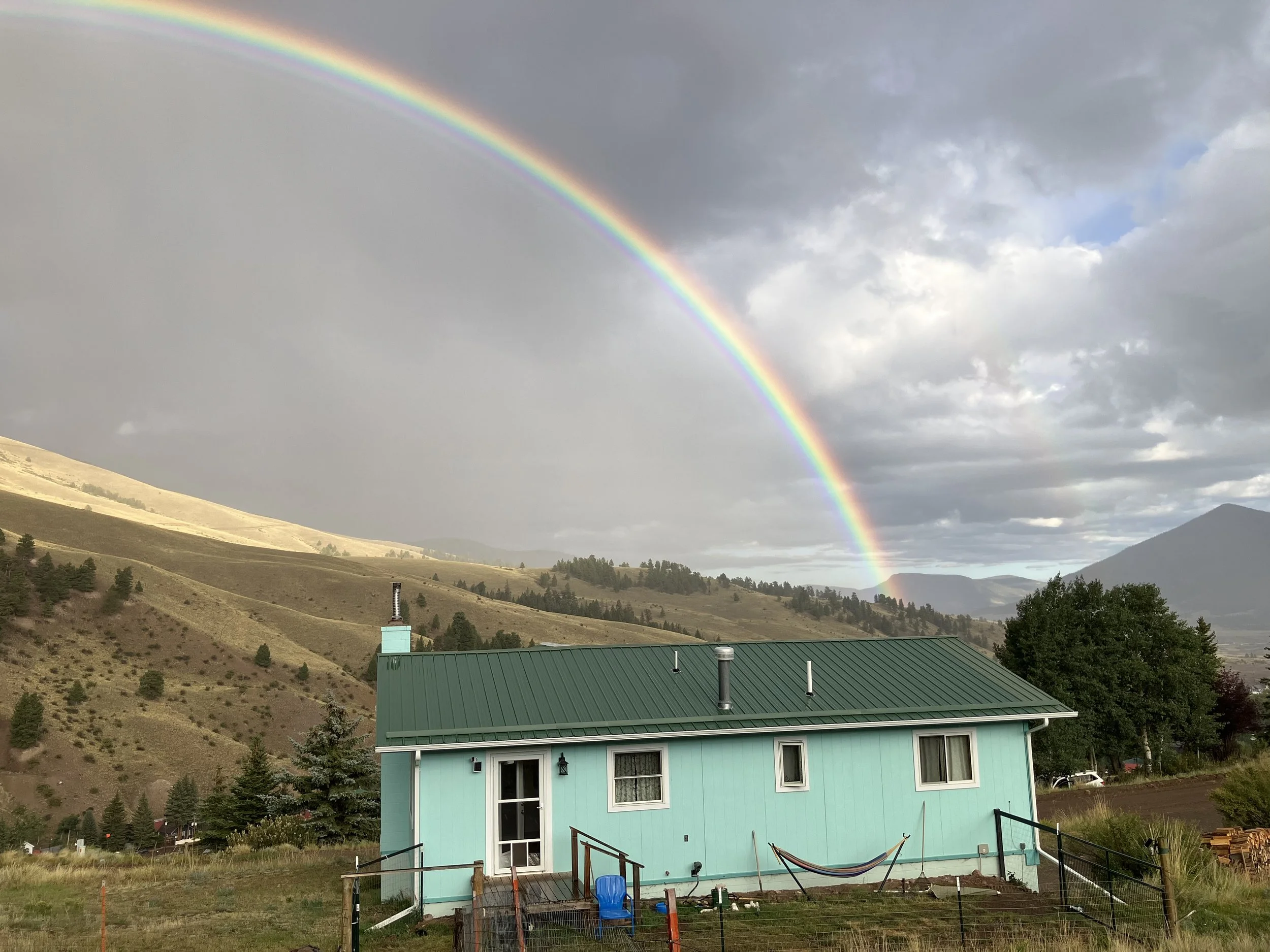 A turquoise house with a green metal roof in a rural landscape under a rainbow and cloudy sky, with hills and trees in the background.