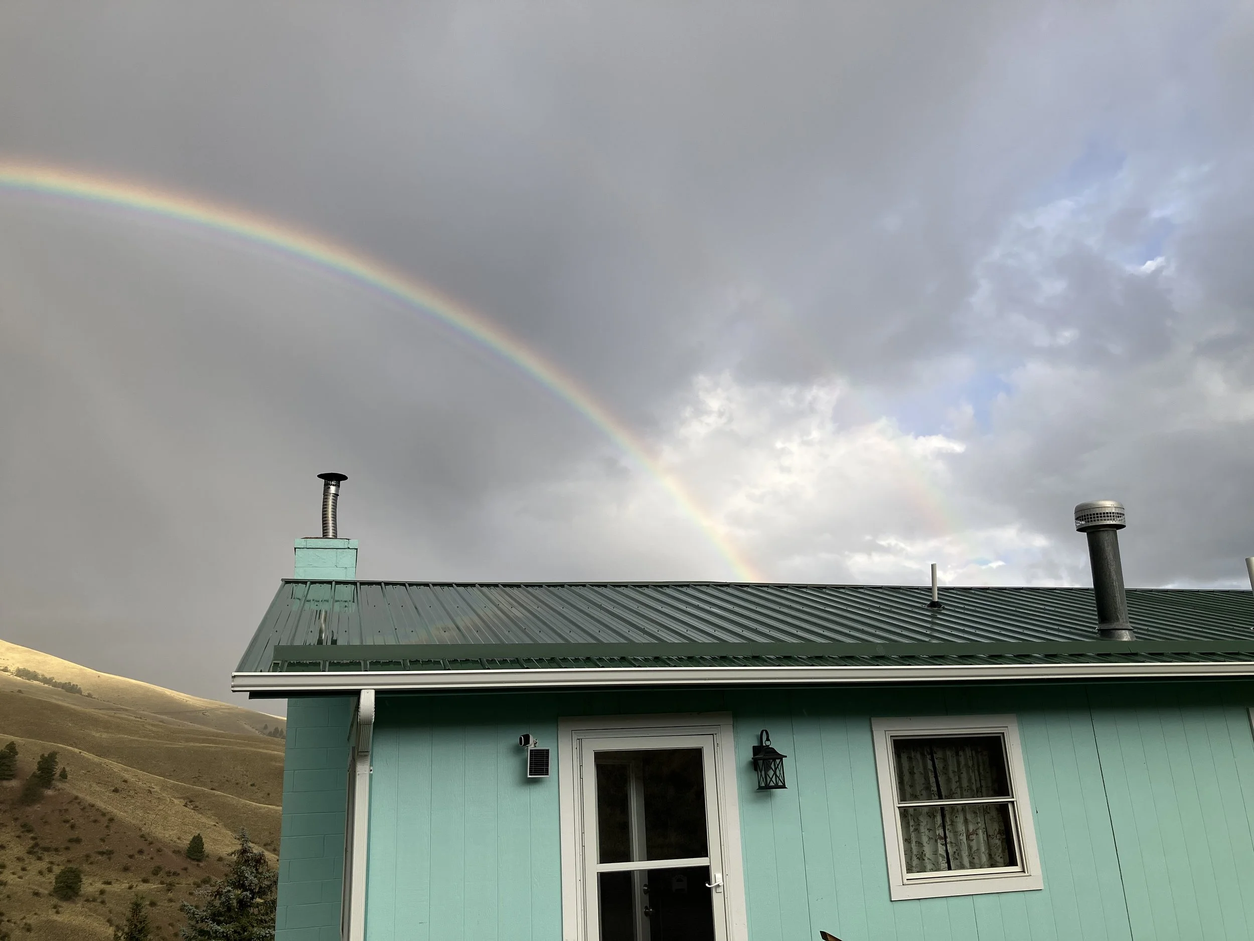 A house with a green metal roof and mint green exterior, two windows, a door, and a lamp outside. A rainbow is visible in the cloudy sky above.