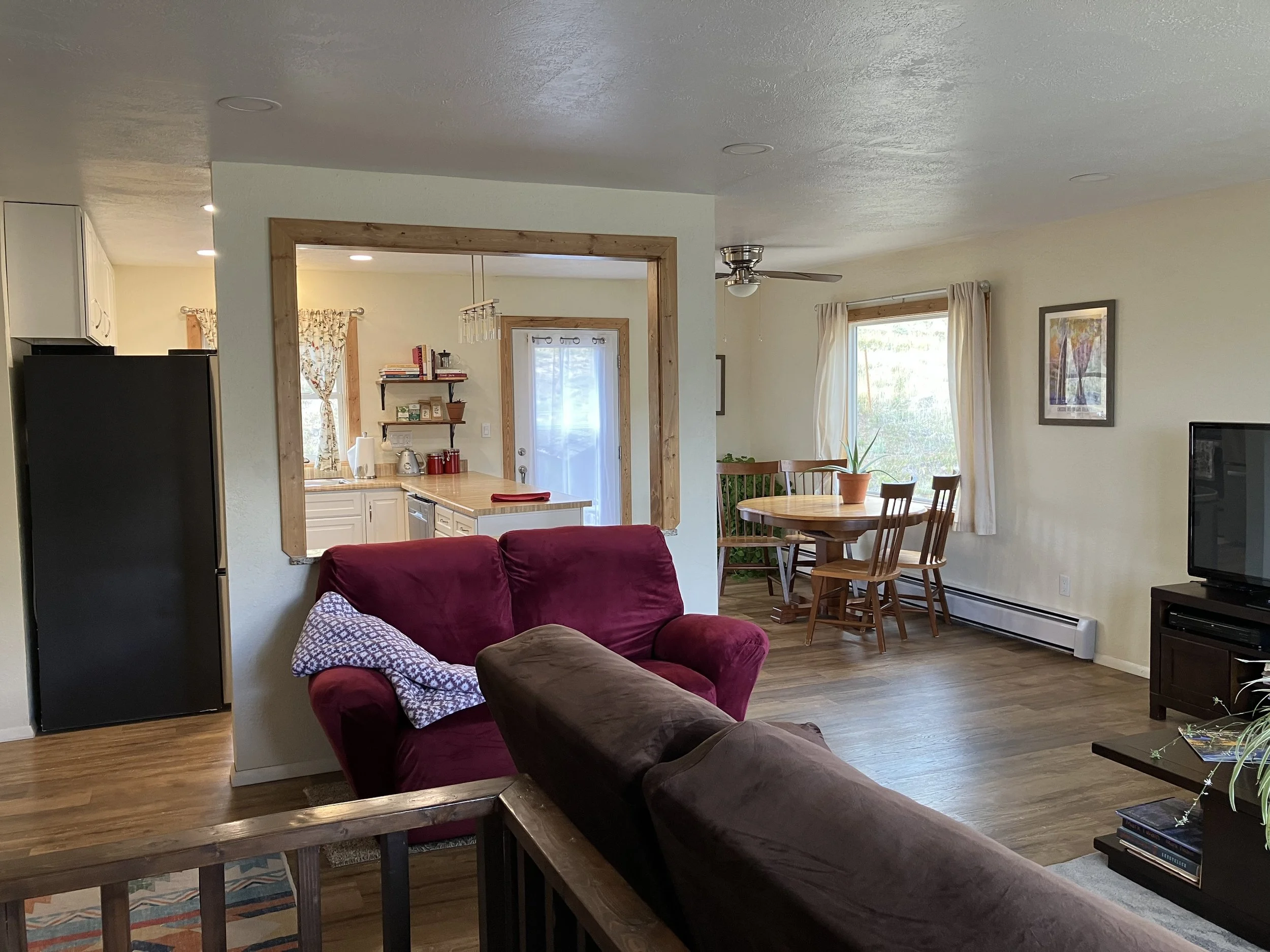 Living room with a brown sofa, a red armchair, a TV, a round wooden dining table with chairs, and a view into the kitchen with white cabinets and a sliding glass door.