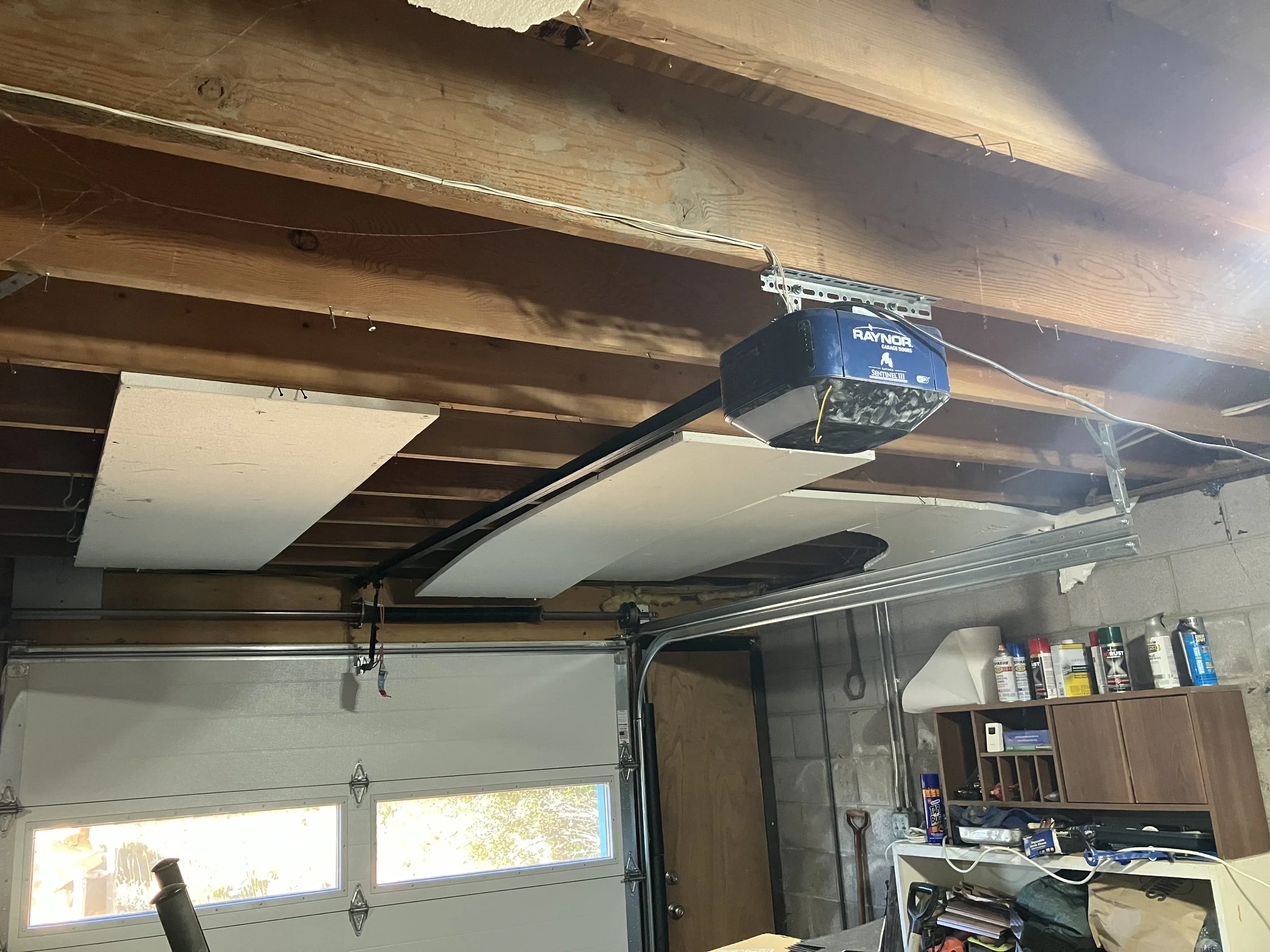 Garage interior with garage door opener, wooden ceiling beams, and a small wooden shelf with cans and tools.