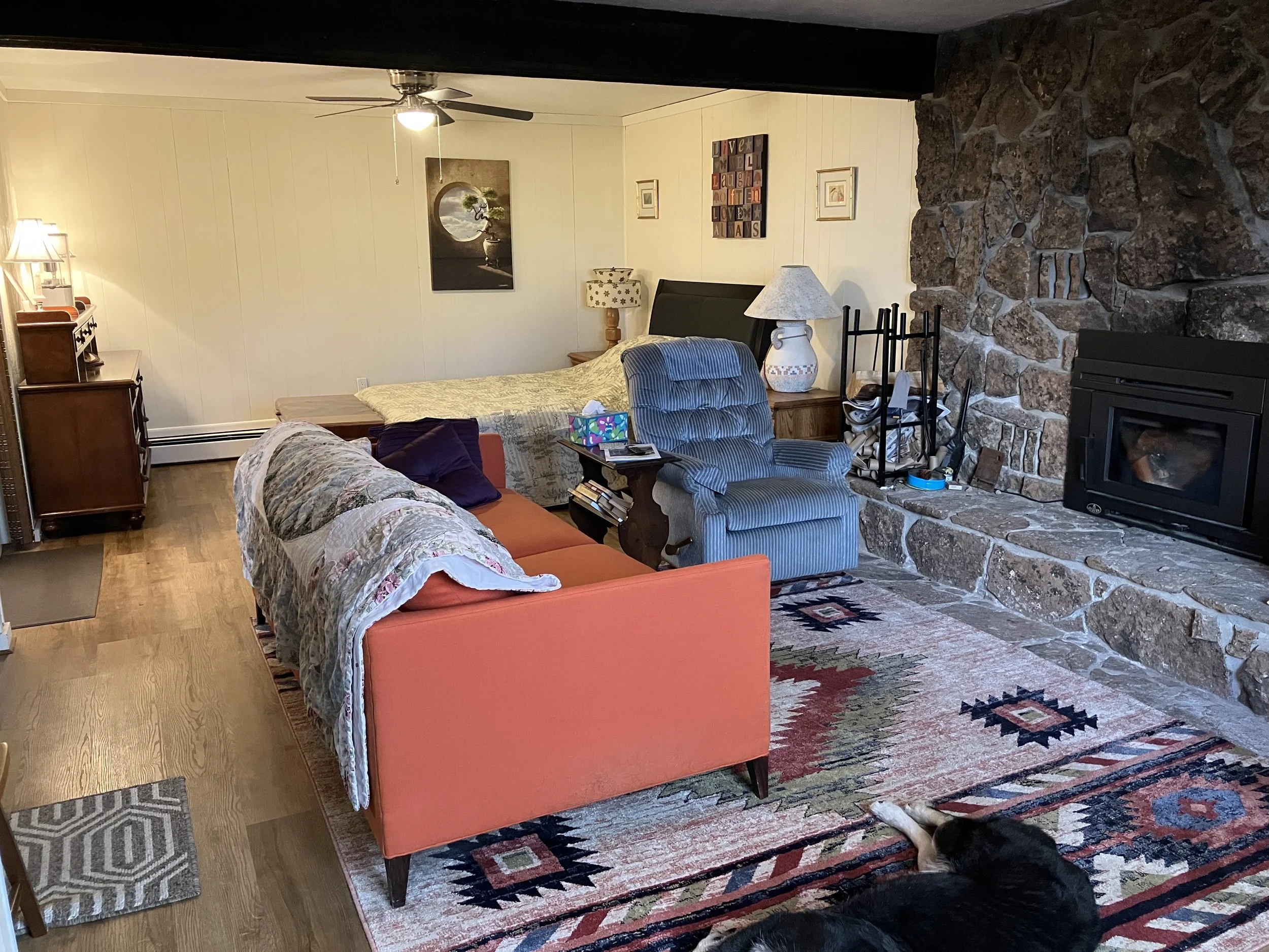 Living room with a stone fireplace, striped armchair, coral sofa with a quilt, and various decorative items. Dog lying on patterned rug.