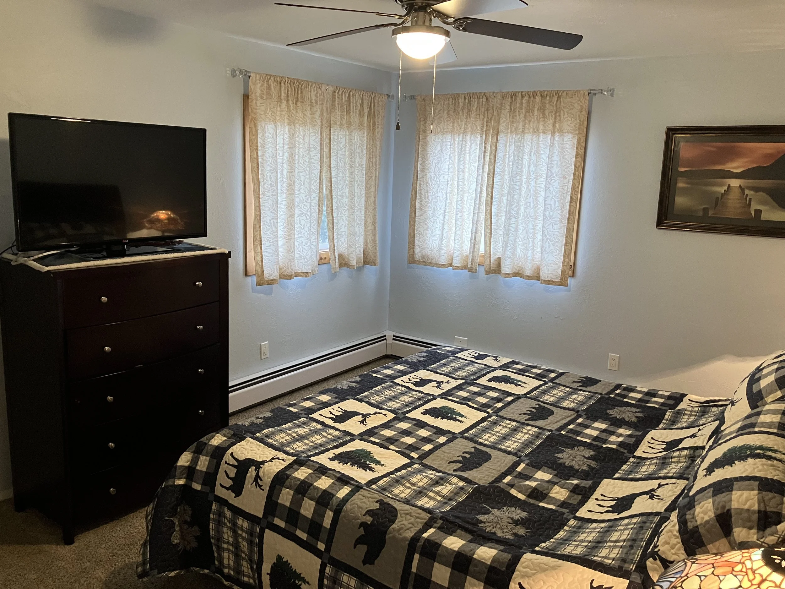 A bedroom with a bed, a dresser with a TV, two windows with beige curtains, and a framed lake landscape painting on the wall.