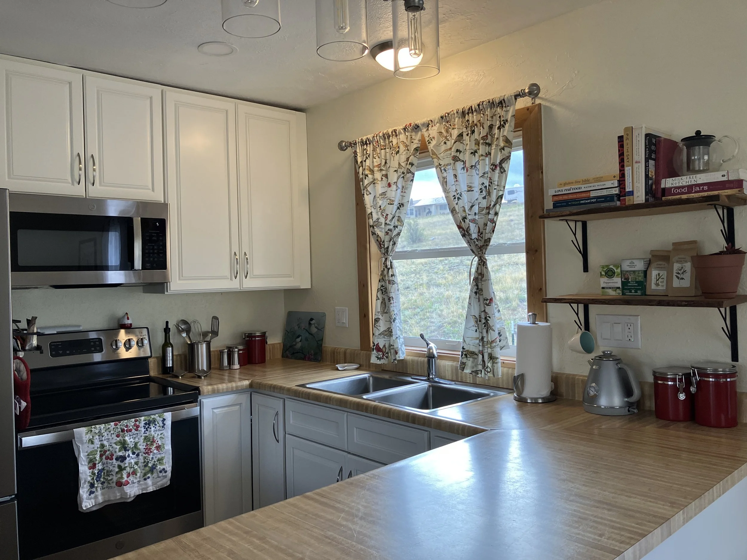 A kitchen with white cabinets, a stainless steel stove, microwave, and a wooden countertop. There are shelves on the wall with books, jars, and a potted plant. A window with floral curtains is above the sink, which has a double basin and a silver fau