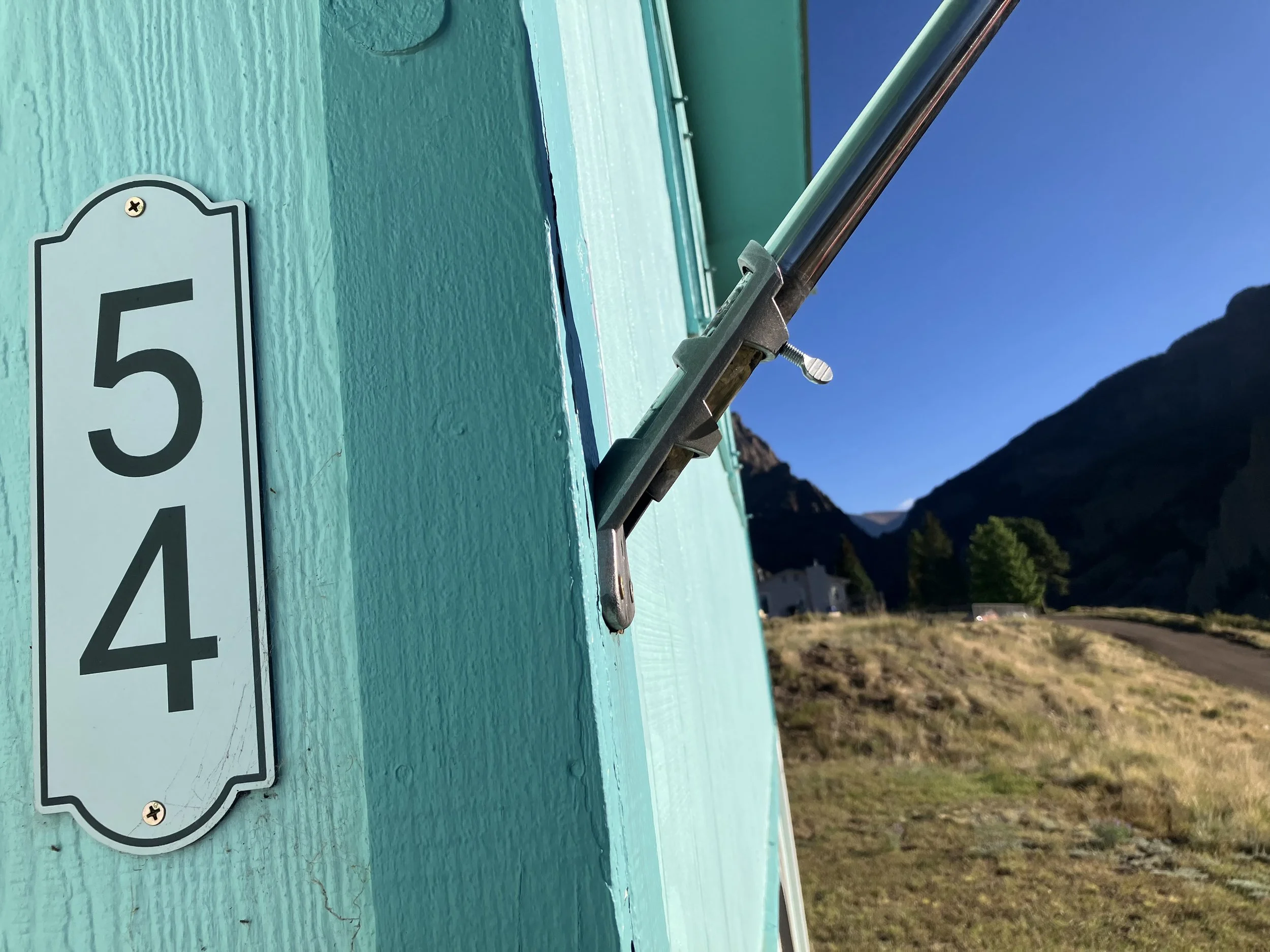 Close-up of a teal-colored mailbox with house numbers 54 on the side, and a postal flag raised, set against a rural landscape with mountains, trees, and a dirt road under a clear blue sky.