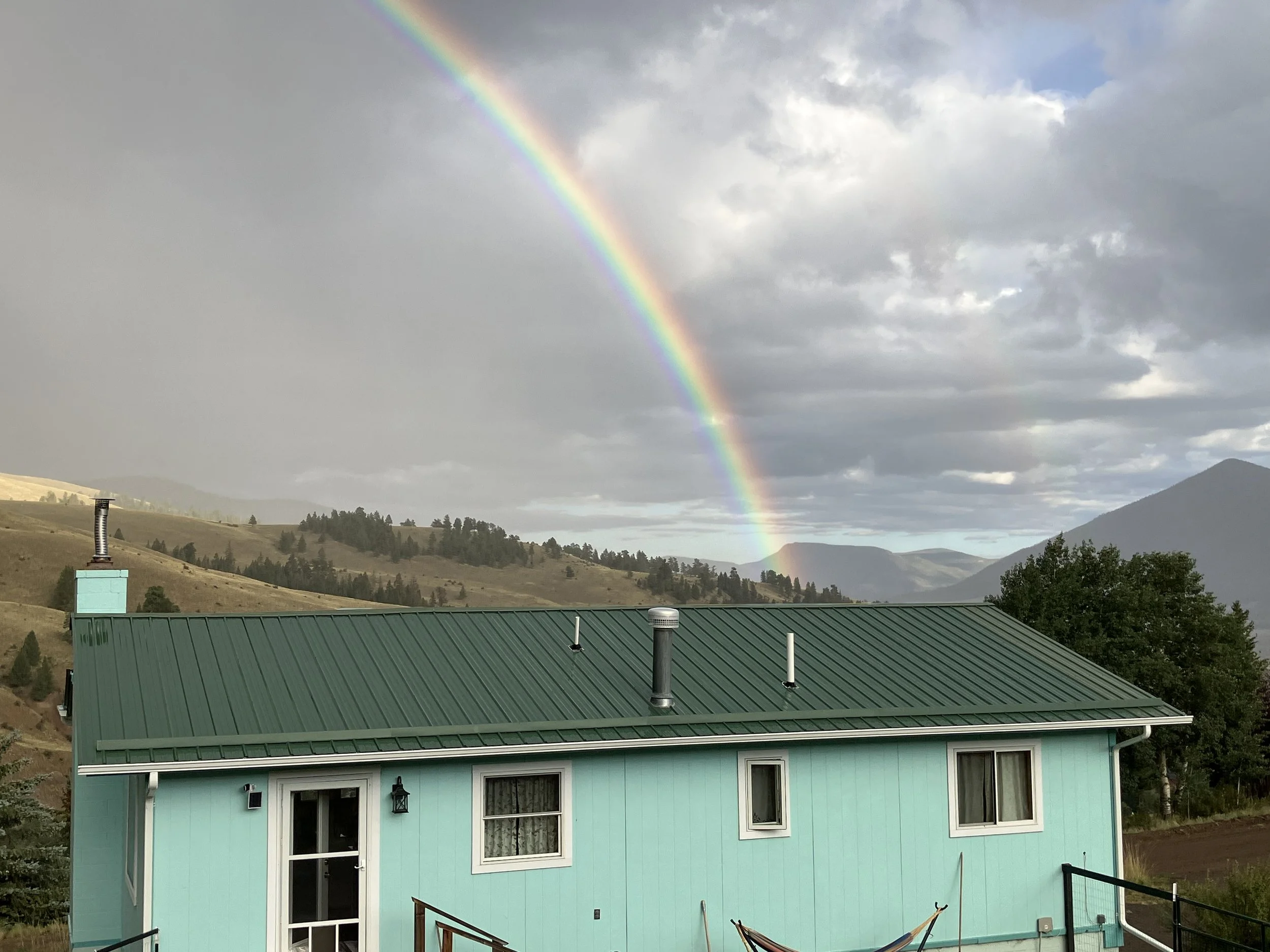 A light blue house with a green metal roof, multiple windows, and a door, with a rainbow in the cloudy sky and rolling hills and mountains in the background.
