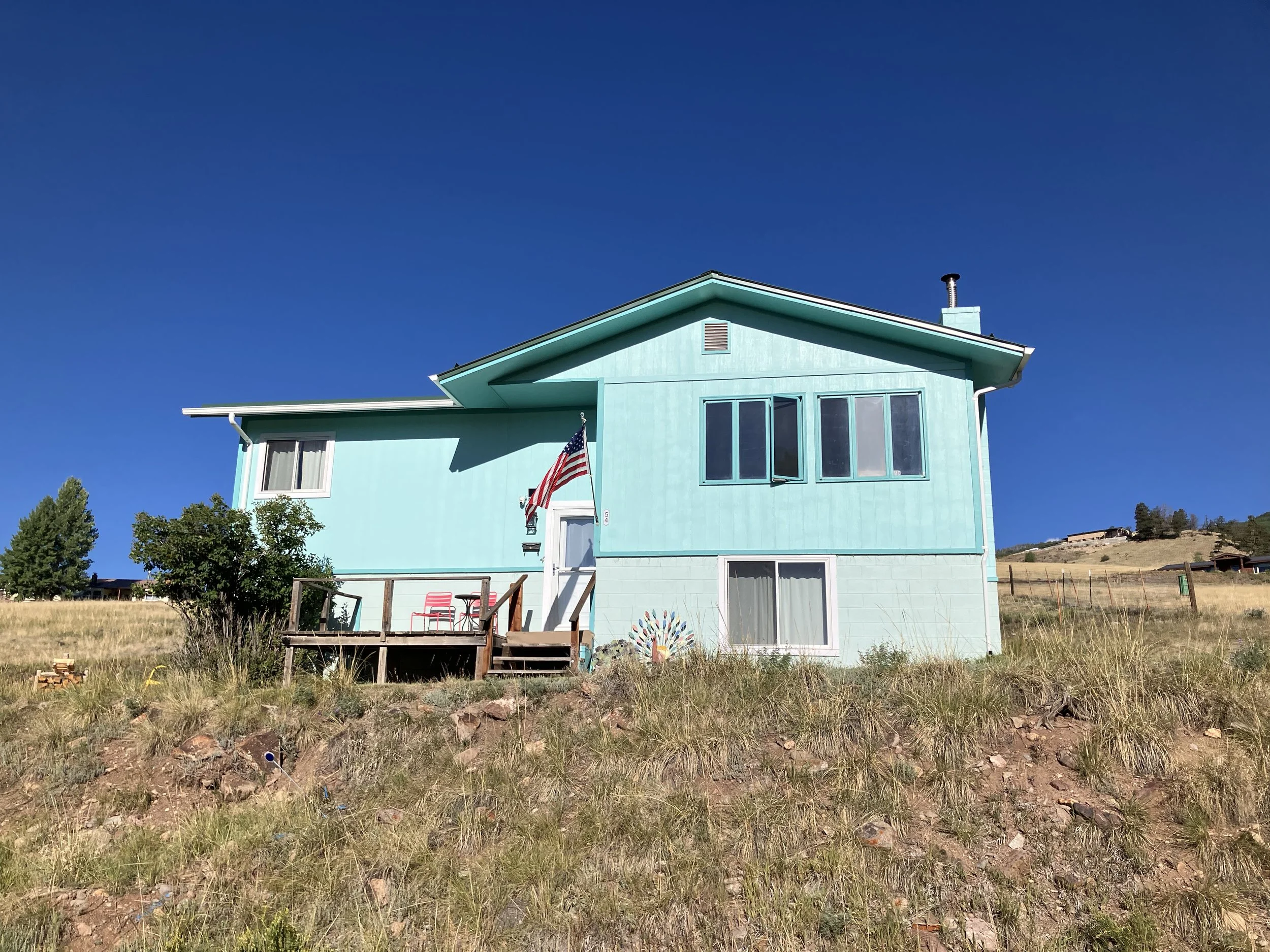 A turquoise two-story house with a small front porch, American flag, and grass surrounding it under a clear blue sky.