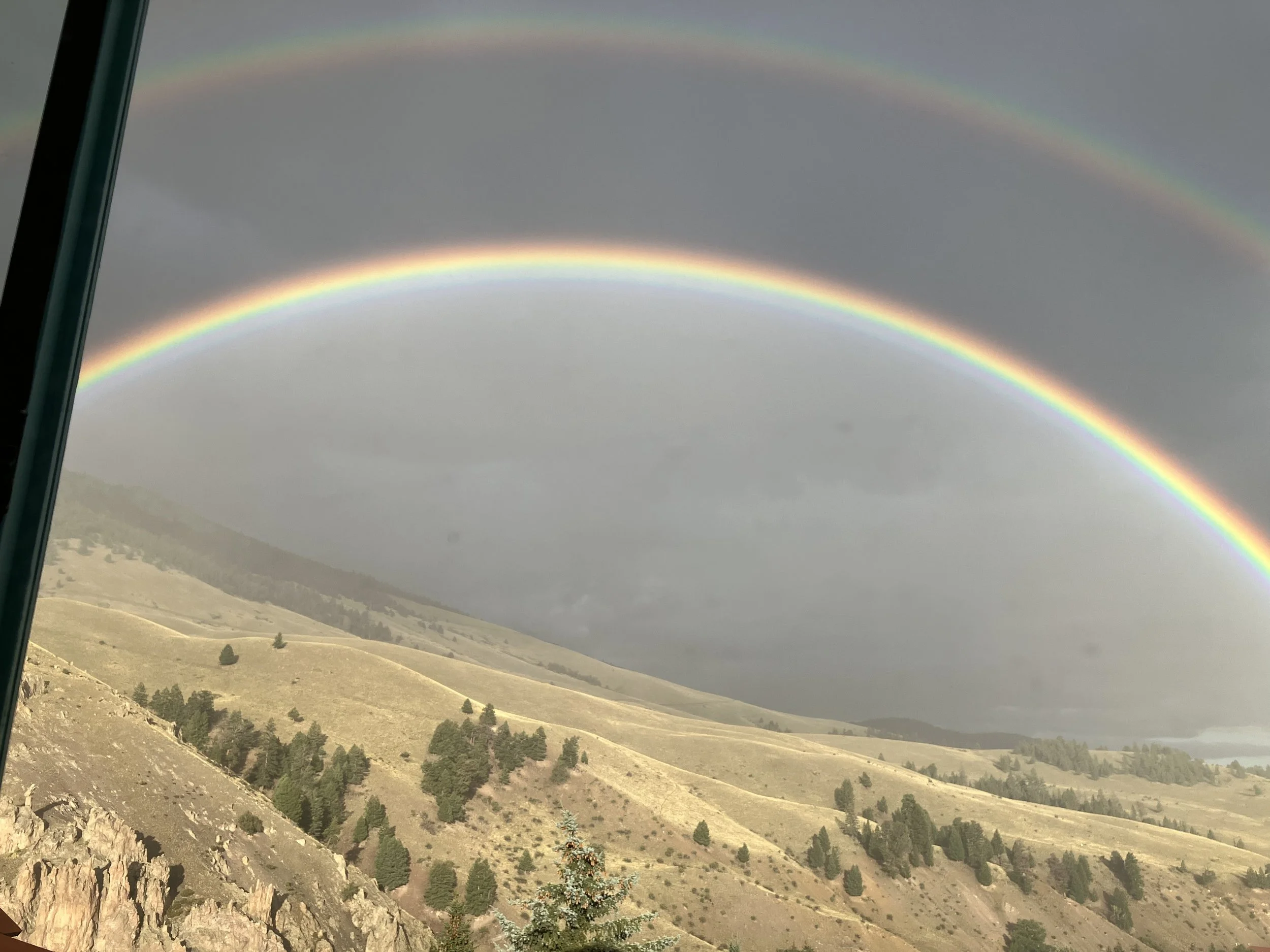 Double rainbow over a mountainous landscape with grassy hills and scattered trees.