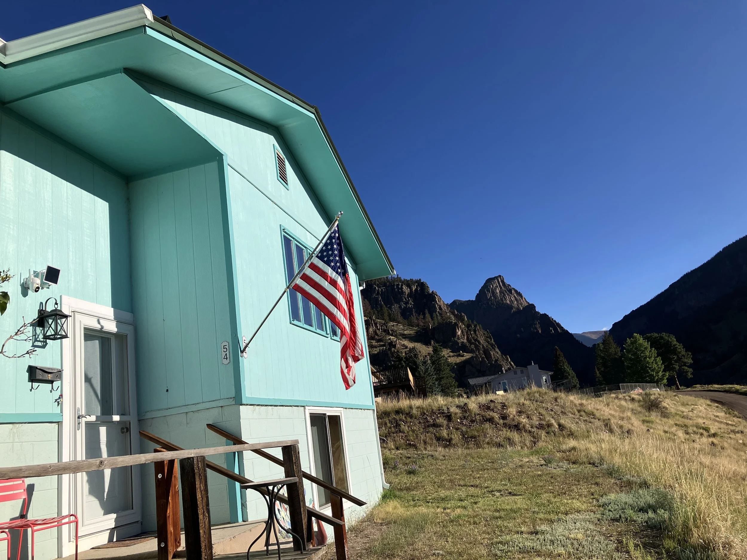 A light blue house with an American flag mounted on the side, situated in a mountainous area with a clear blue sky.