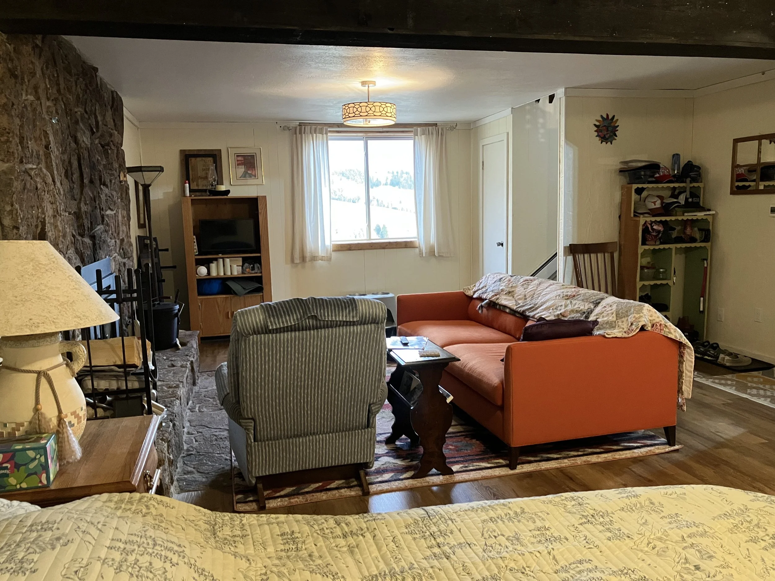 Living room with stone fireplace, orange couch, green striped armchair, TV stand, window with curtains, and various decorative items.