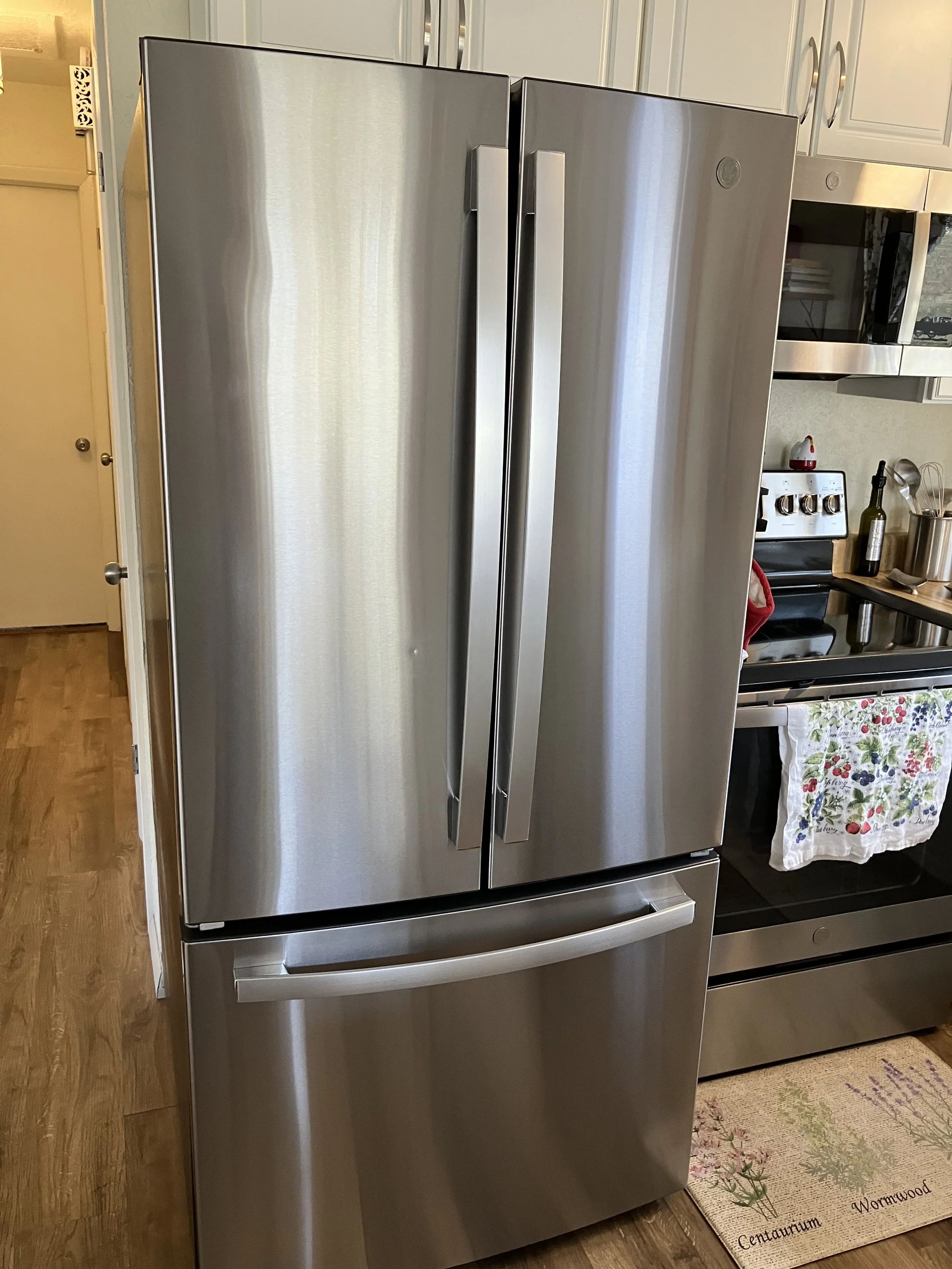 Stainless steel refrigerator in a kitchen with a double door freezer and a bottom pull-out drawer, next to a stove with a floral dish towel hanging on the oven handle.