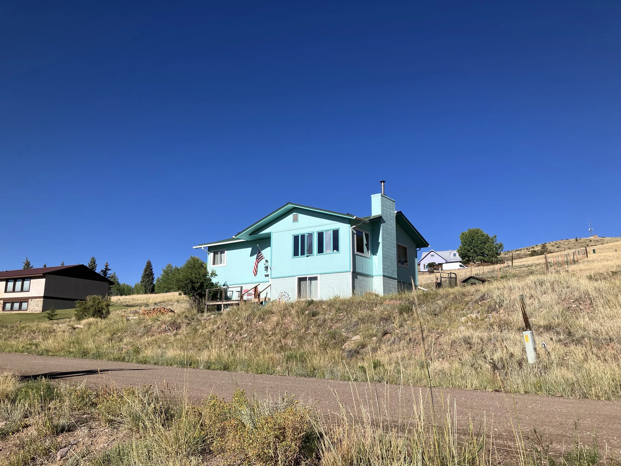 A light blue house on a grassy hillside with a clear blue sky above. There are other houses in the background and an American flag hanging on the house.