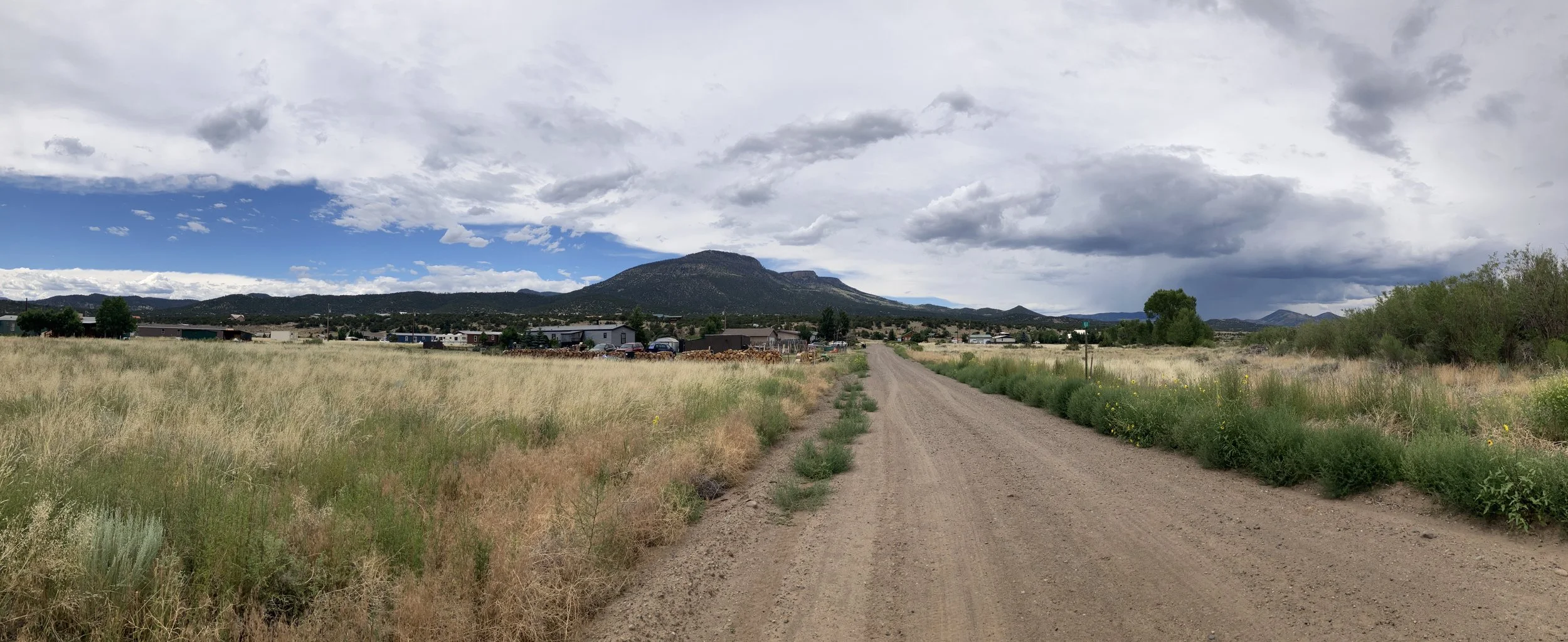 Dirt road leading towards houses and a mountain in the background under partly cloudy sky
