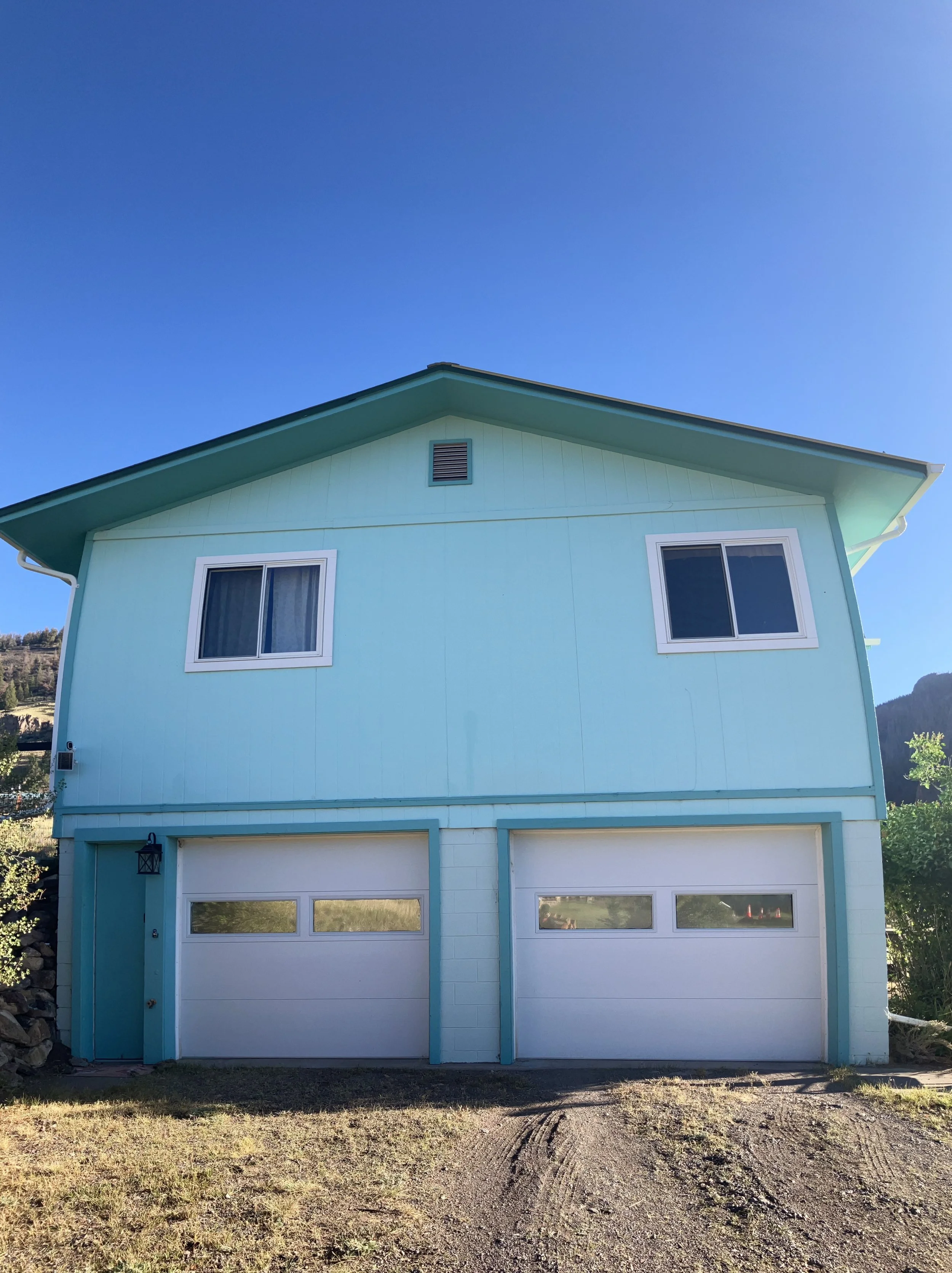 A two-story house painted in light blue with white garage doors, two windows on the upper floor, and a small vent under the roof. The house has a gravel driveway and is set against a hilly landscape under a clear blue sky.