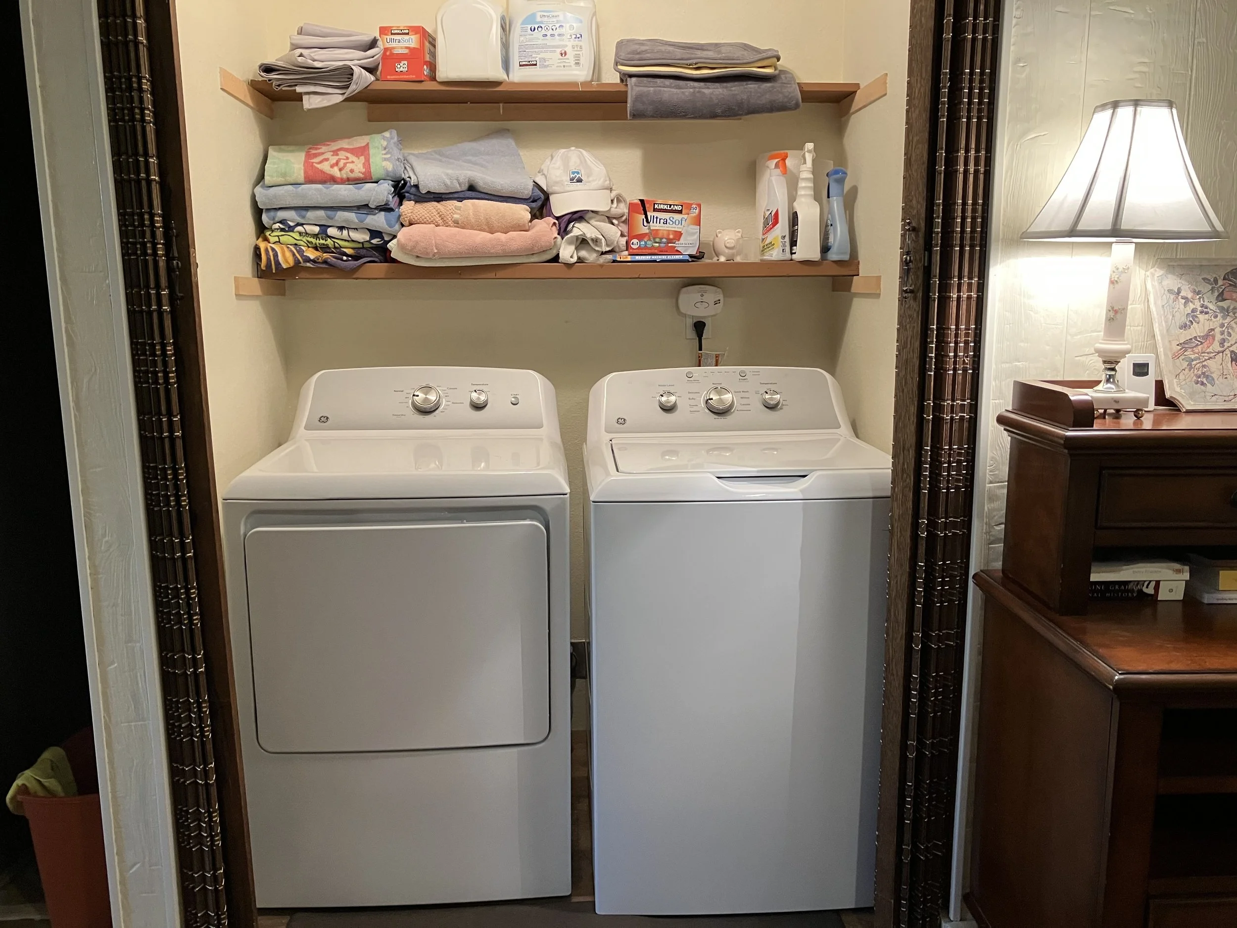 Laundry room with a white washing machine and dryer, two brown wooden shelves above with folded towels, cleaning supplies, and laundry detergent. A lamp and wooden cabinet are on the right side.
