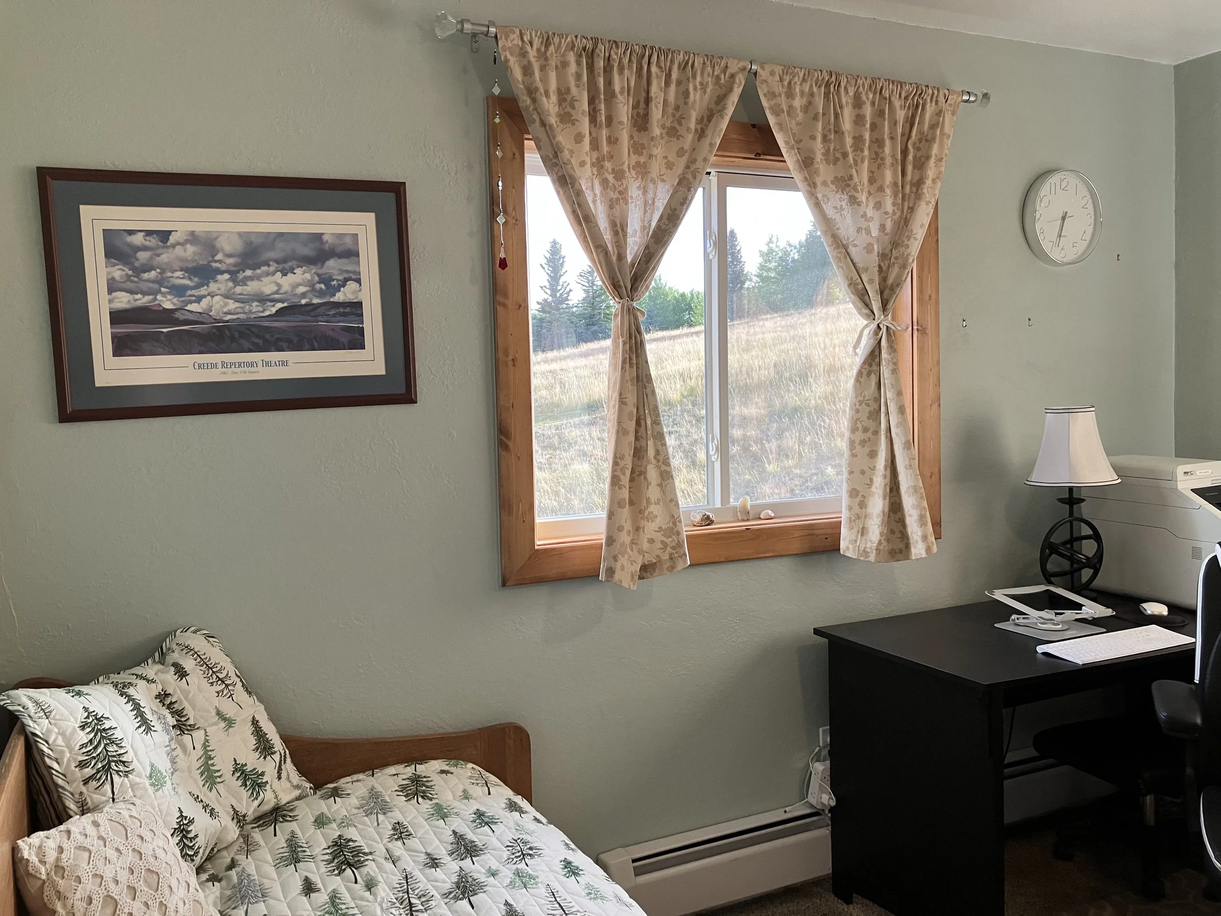 A cozy room with a bed that has a forest-themed quilt, a wooden window with floral curtains, a framed landscape picture on the wall, a black desk with a computer, a white lamp, and a wall clock showing 10:07.
