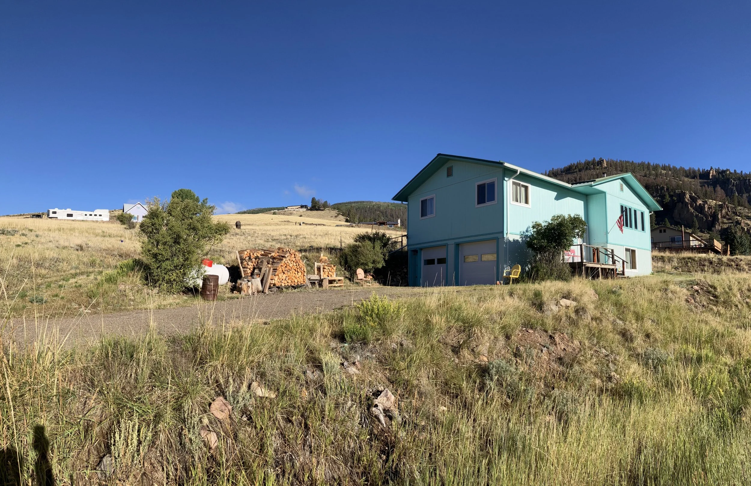 A blue two-story house with a garage and an American flag on the porch, situated on a grassy hillside with a tree and woodpile, under a clear blue sky.