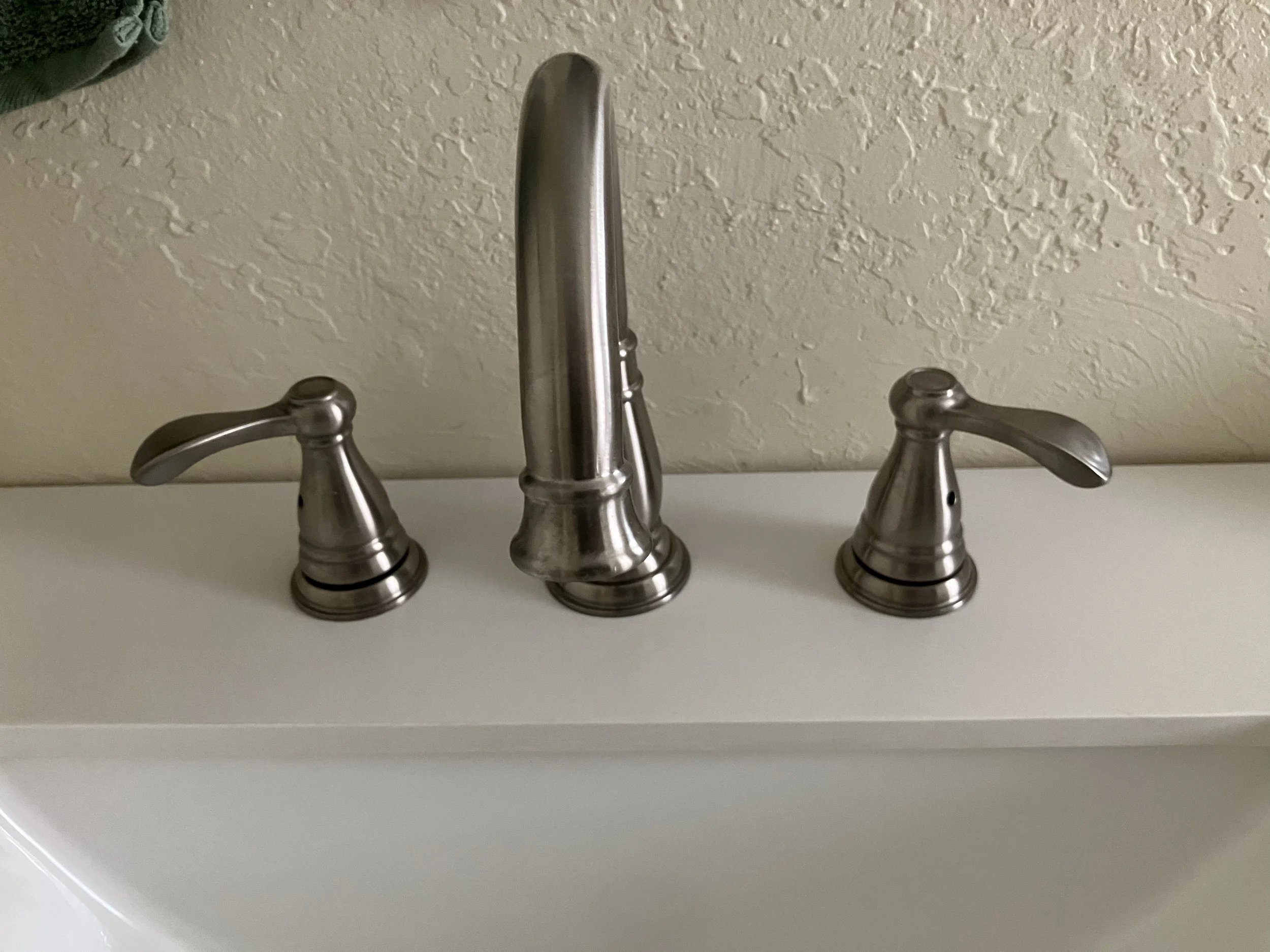 A brushed nickel bathroom faucet with two handles, mounted on a white sink against a textured beige wall.
