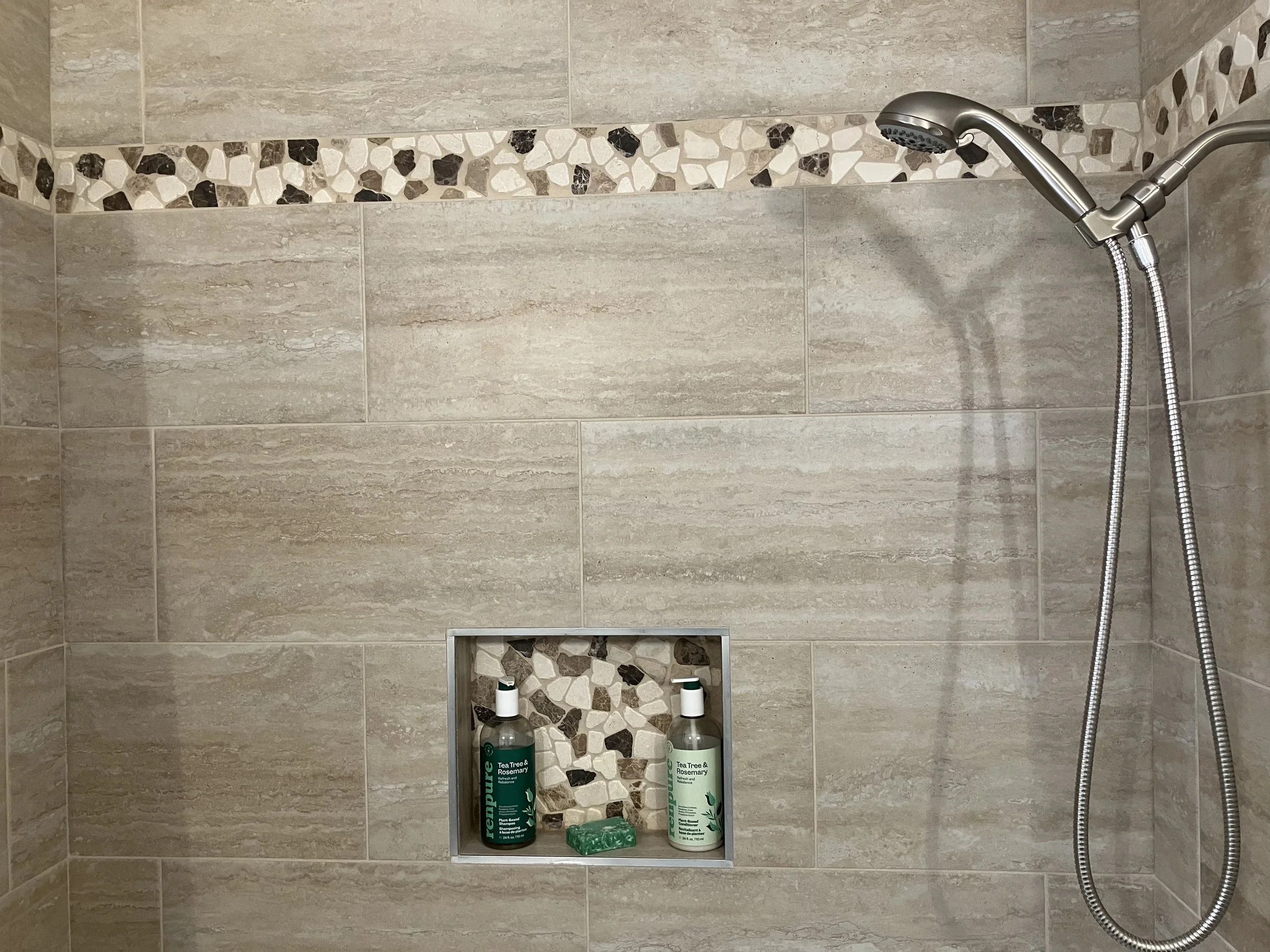 Shower with two green bottles of tea tree and rosemary shampoo and conditioner, and soap in a recessed shelf, on beige tiled wall with mosaic accents.