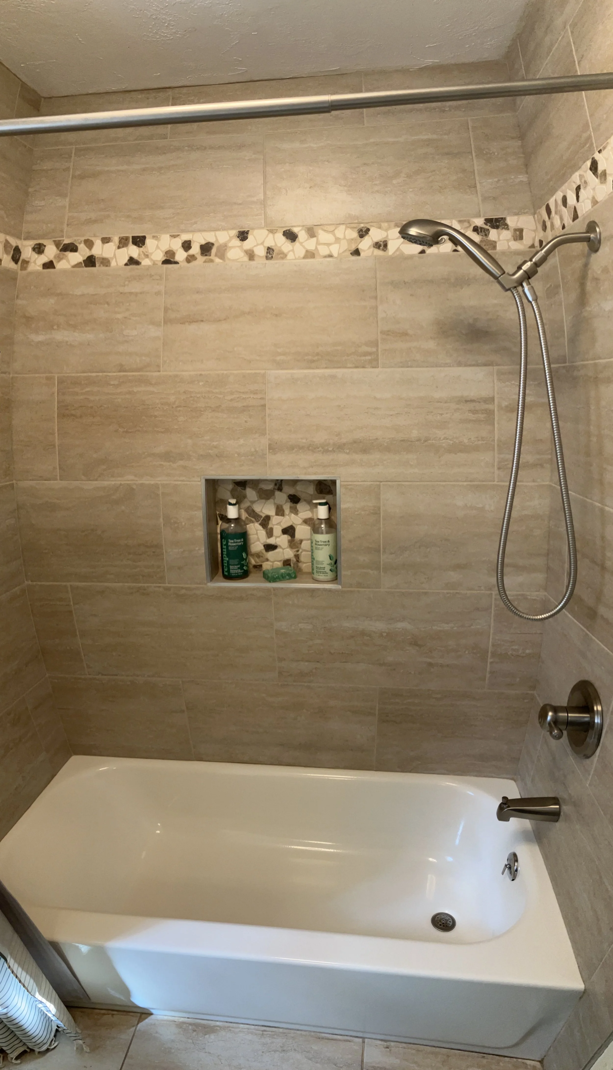 A bathroom shower with beige tiles, a built-in niche with shampoo bottles, a handheld showerhead, and a white bathtub.