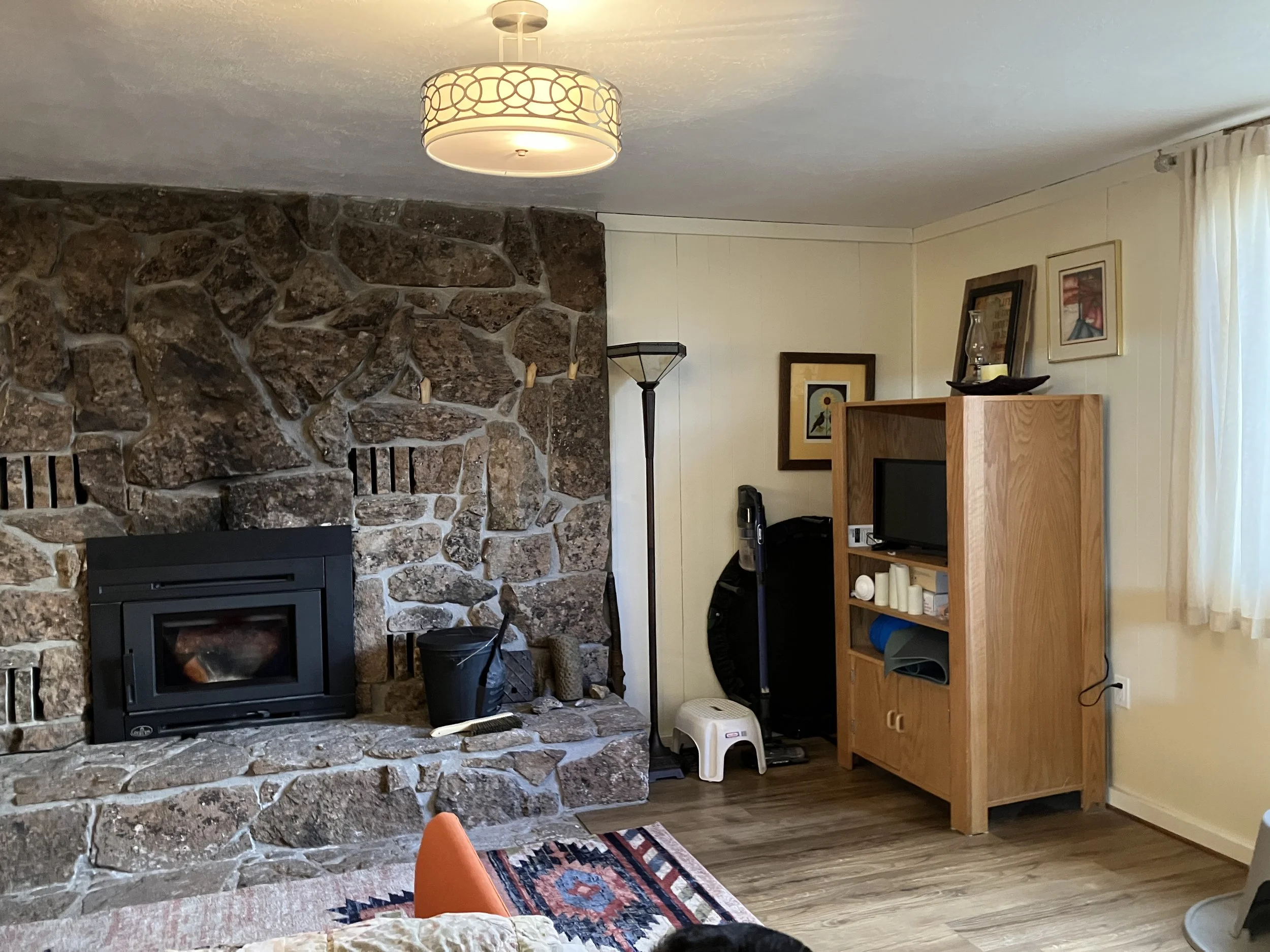 Living room with a stone fireplace, a hanging ceiling light, a wooden shelving unit with a small TV, candles, and framed artwork, a floor lamp, window with white curtains, and various household items.