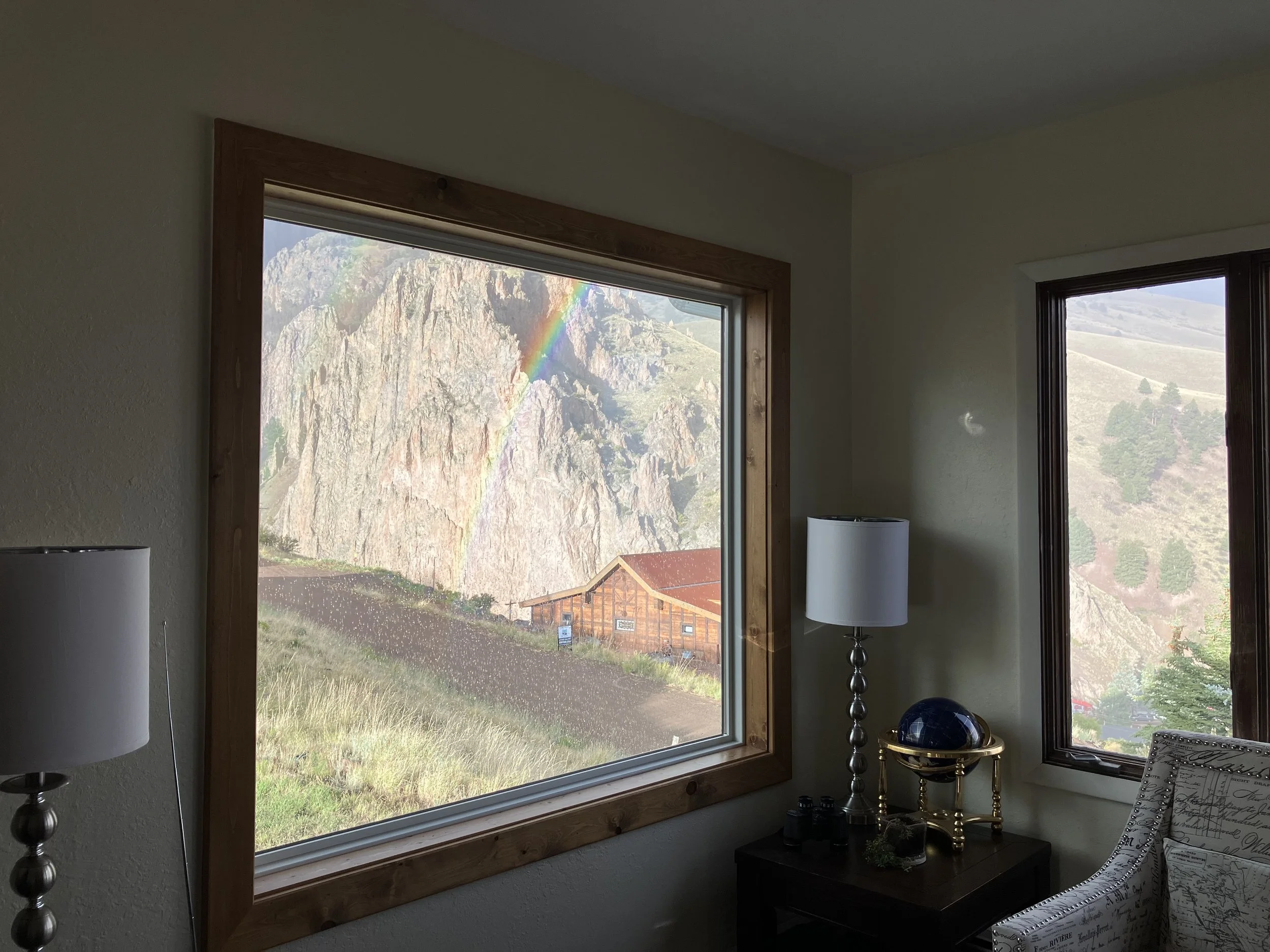 Room with large window showing a mountain landscape, rainbow, and building outside, interior decor includes lamps, a table with a globe, and patterned armchair.
