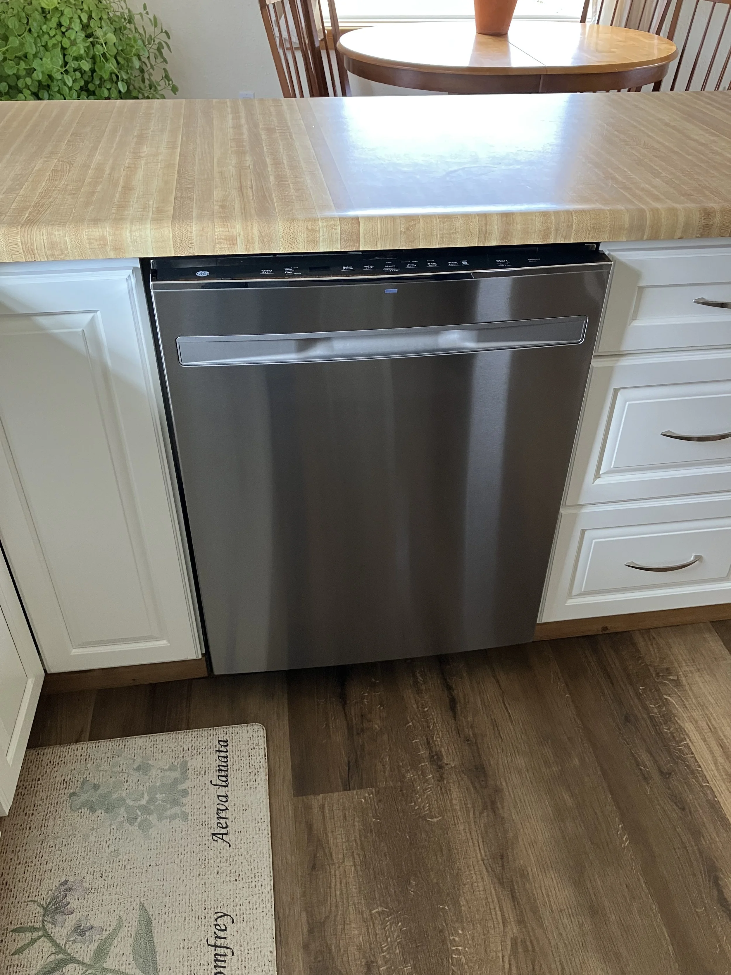 Stainless steel dishwasher installed below a wooden kitchen countertop with white cabinetry on either side. A round wooden dining table and chairs are visible in the background.
