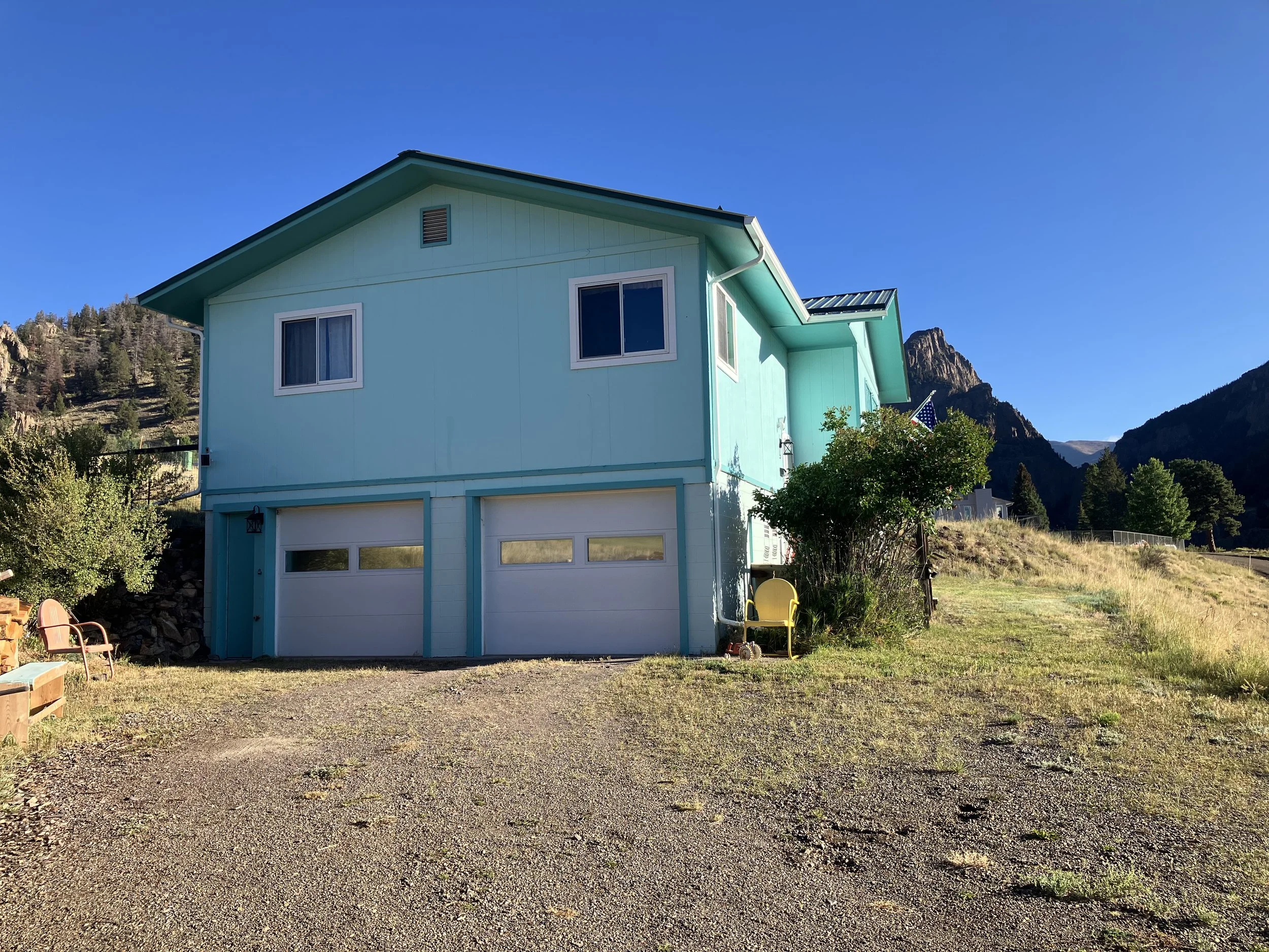 A two-story light blue house with a garage in a rural area surrounded by mountains and trees, under a clear blue sky.