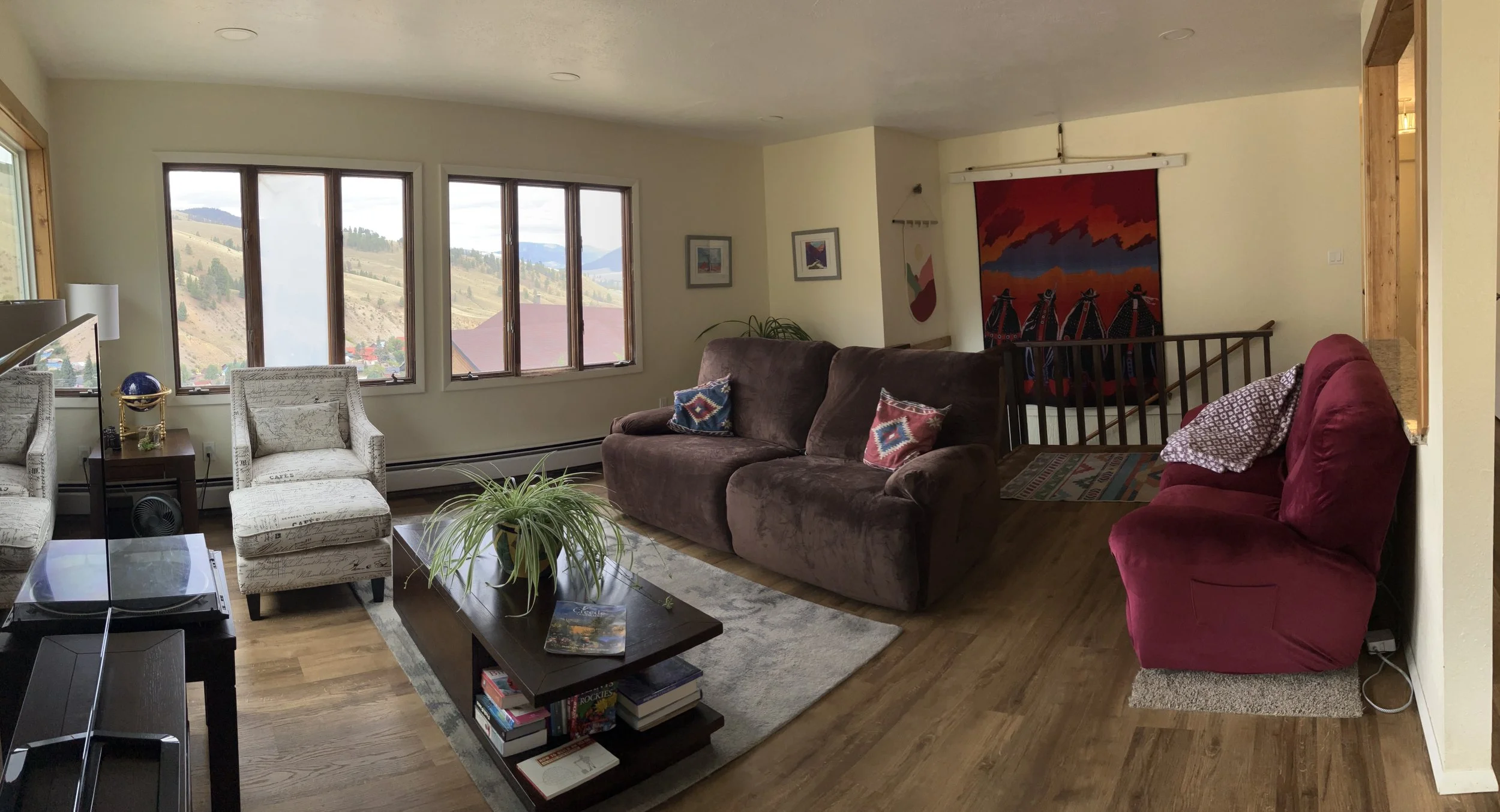 Living room with large windows overlooking hilly landscape, beige walls, dark wooden floors, various chairs including a white armchair and a red armchair, brown sofa with decorative pillows, wooden coffee table with books and a potted plant, wall art