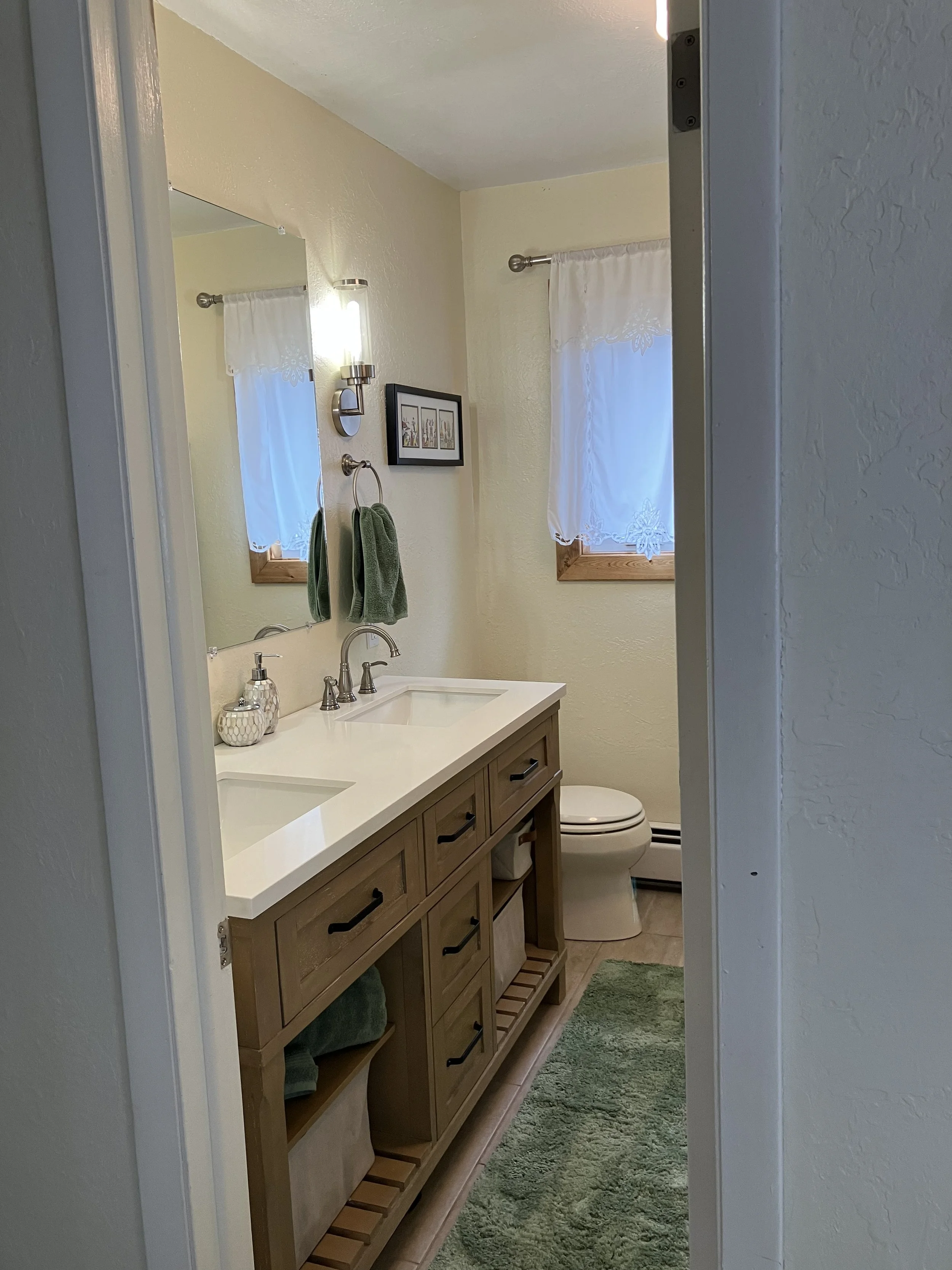 A small bathroom with a double sink vanity, a mirror, a wall light, a small framed picture, two small windows with white curtains, a green towel on a ring, a green rug, and a white toilet.