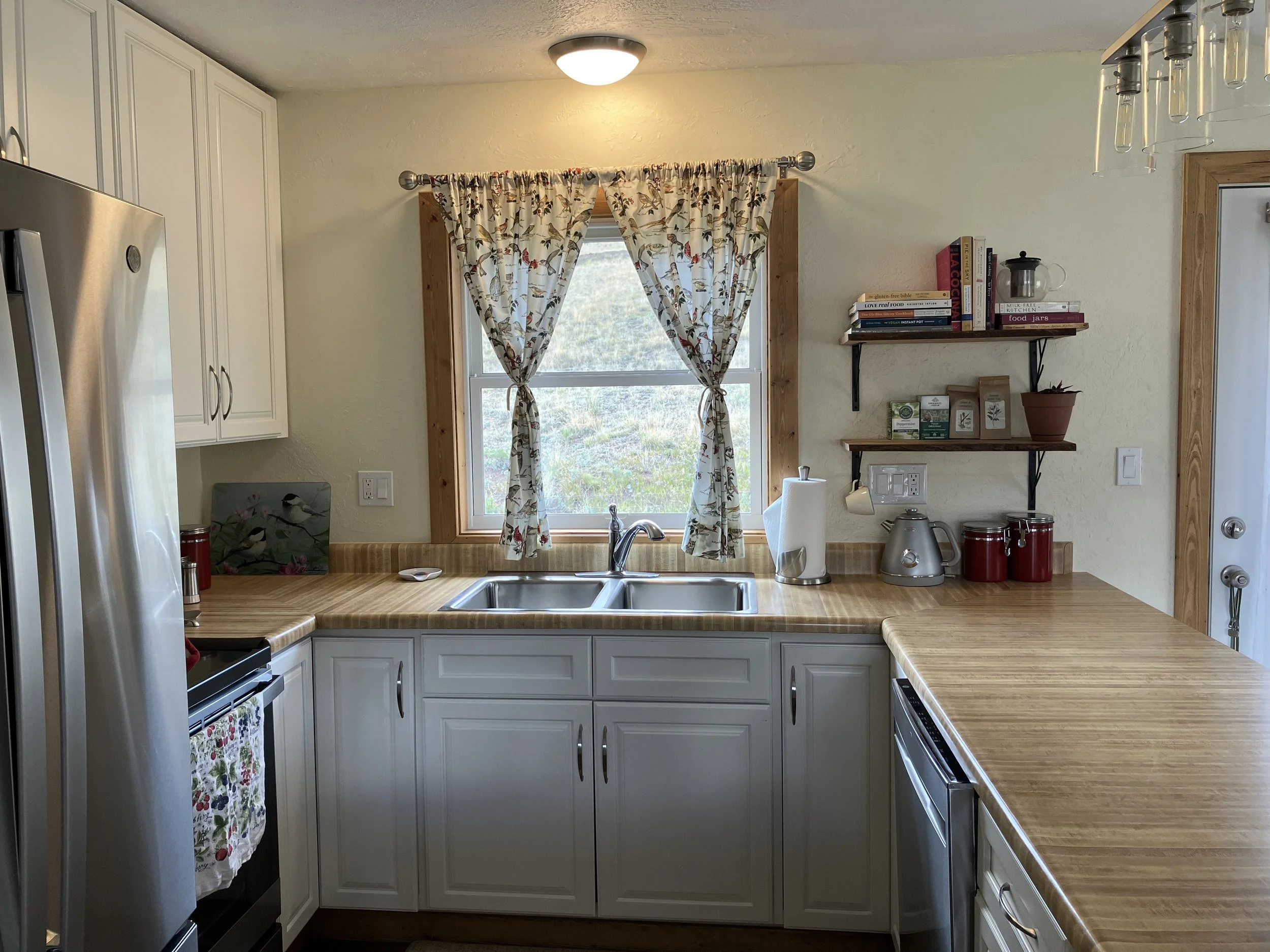 Kitchen with white cabinets, wood countertops, stainless steel refrigerator, stove, and sink. Window with floral curtains above the sink, open to a view of outdoor greenery.