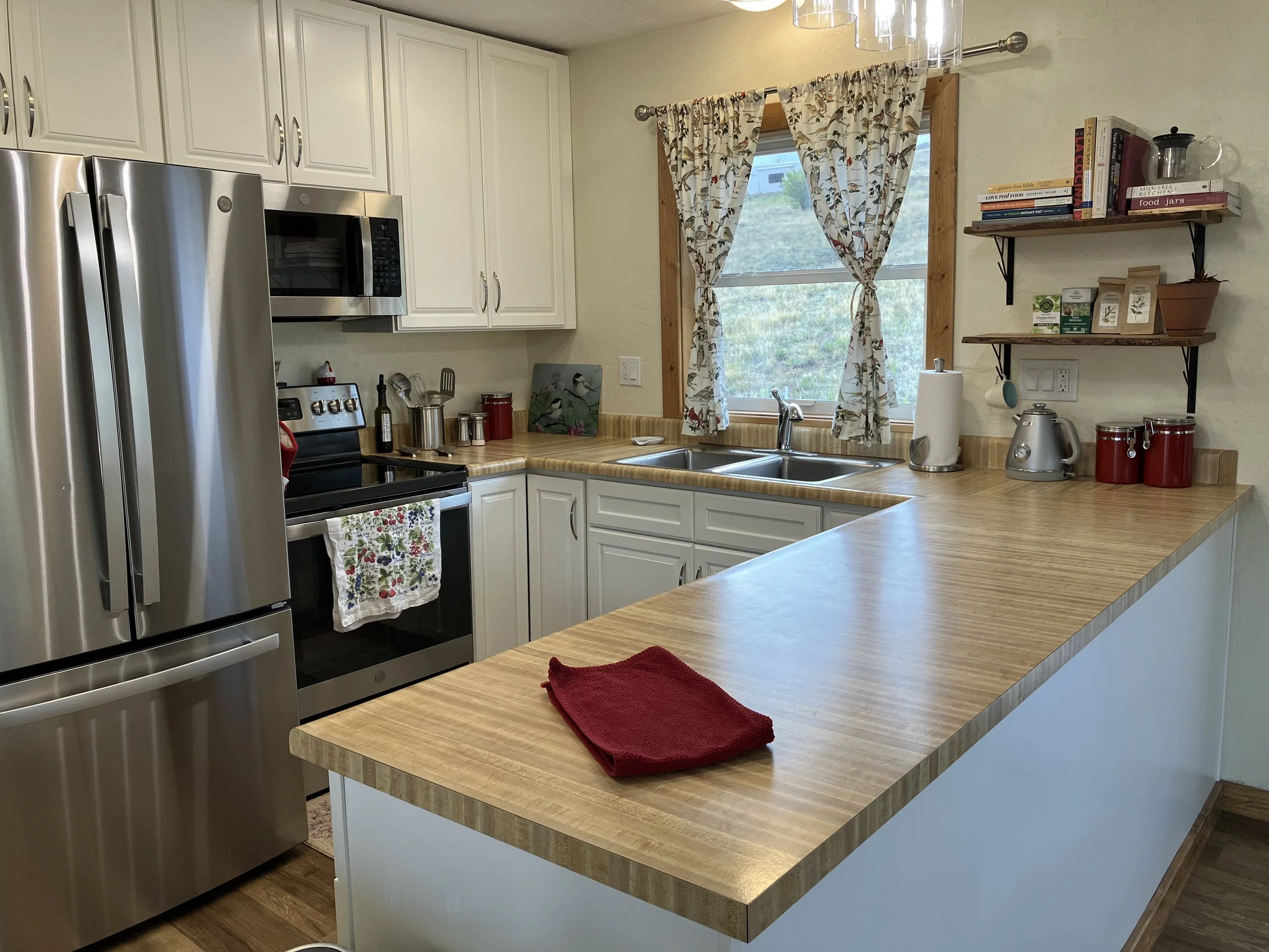 Kitchen with white cabinets, stainless steel refrigerator, stove, microwave, and a window with floral curtains. Wooden countertops with a red cloth, paper towel roll, and kitchen appliances. Open shelving with books, jars, and potted plants.
