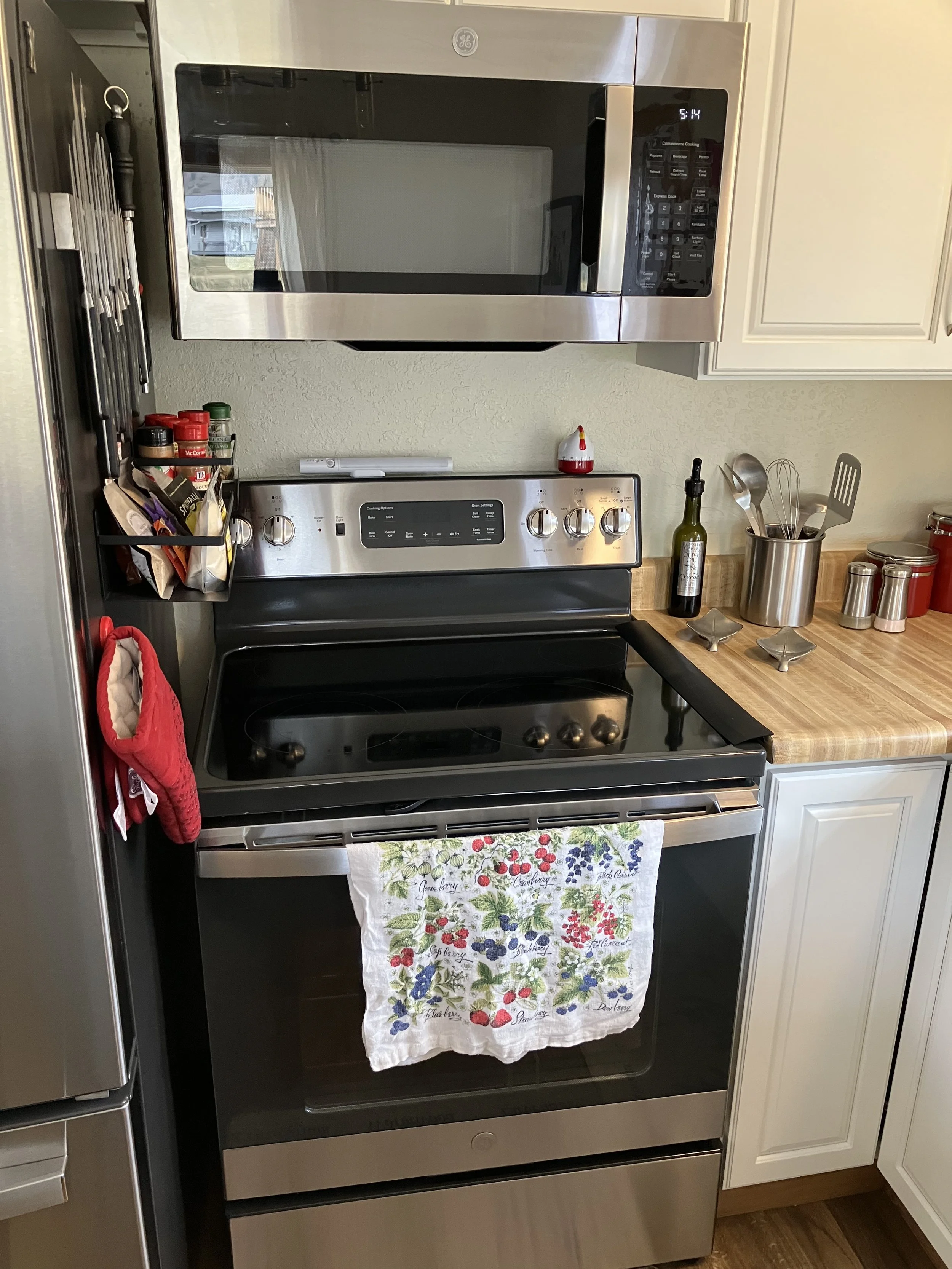 A kitchen with a stainless steel microwave installed above a stainless steel electric stove. The stove has a colorful towel with berry illustrations hanging on the oven handle. To the right of the stove is a wooden countertop with a metal utensil hol