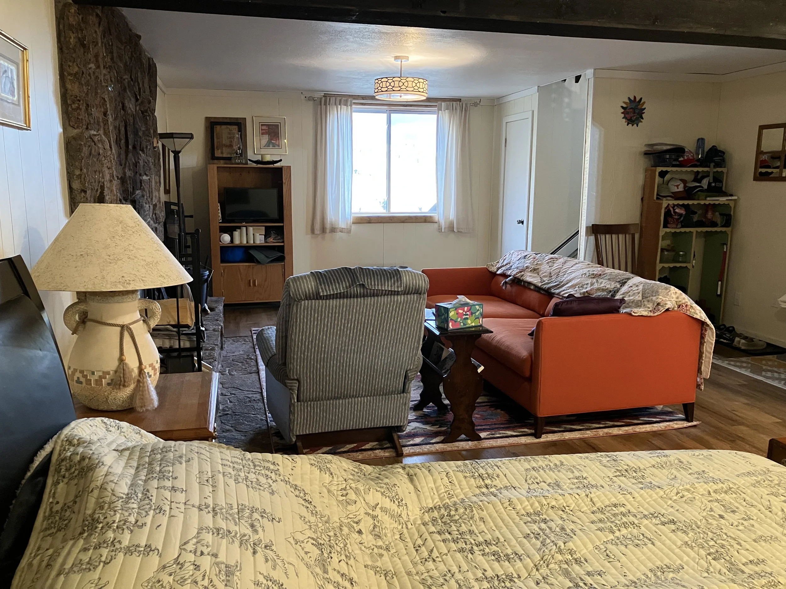 A cozy living room with a bed in the foreground, featuring a patterned beige quilt. To the left, a lamp with a tassel decorates a wooden side table. A stone fireplace is on the left wall. In the center, there are two chairs—one striped and one orange